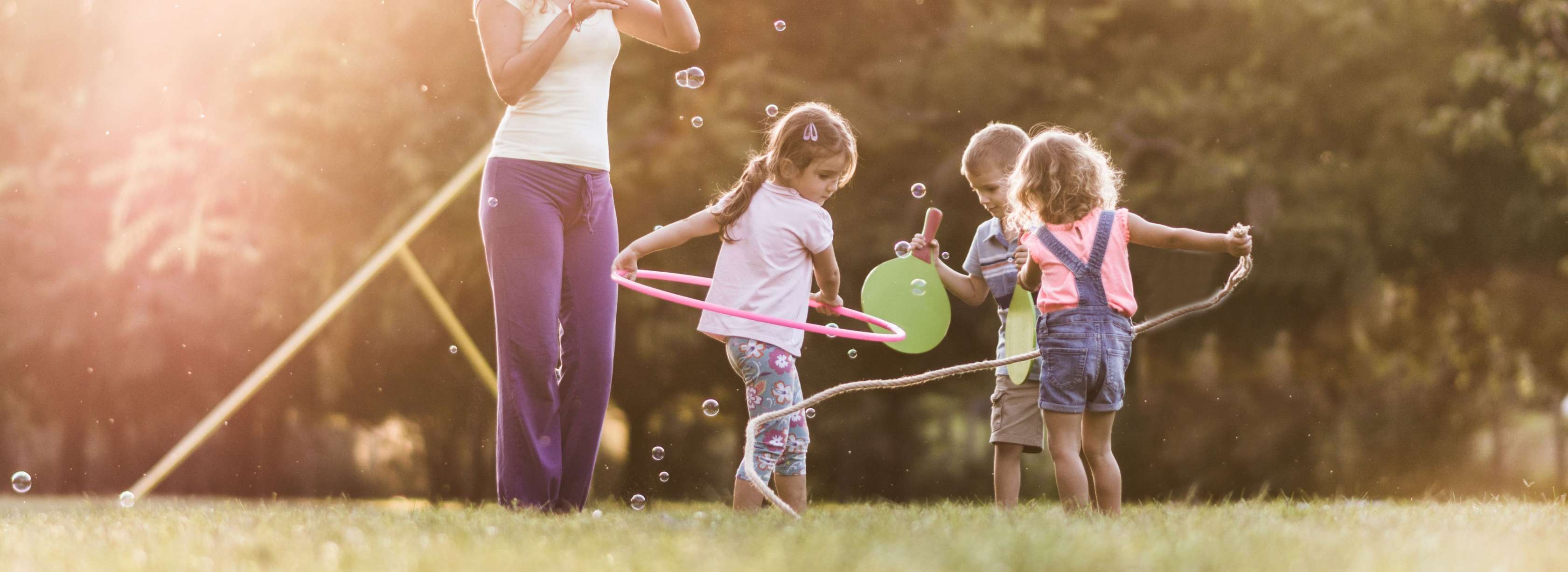 Eine junge Kindergärtnerin steht mit drei spielenden Kindern im Garten und bläst Seifenblasen.