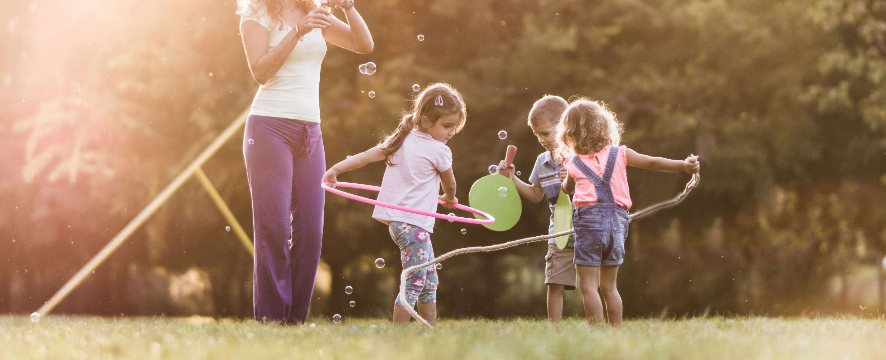 Allianz - Kita gründen | Eine junge Kindergärtnerin steht mit drei spielenden Kindern im Garten und bläst Seifenblasen.