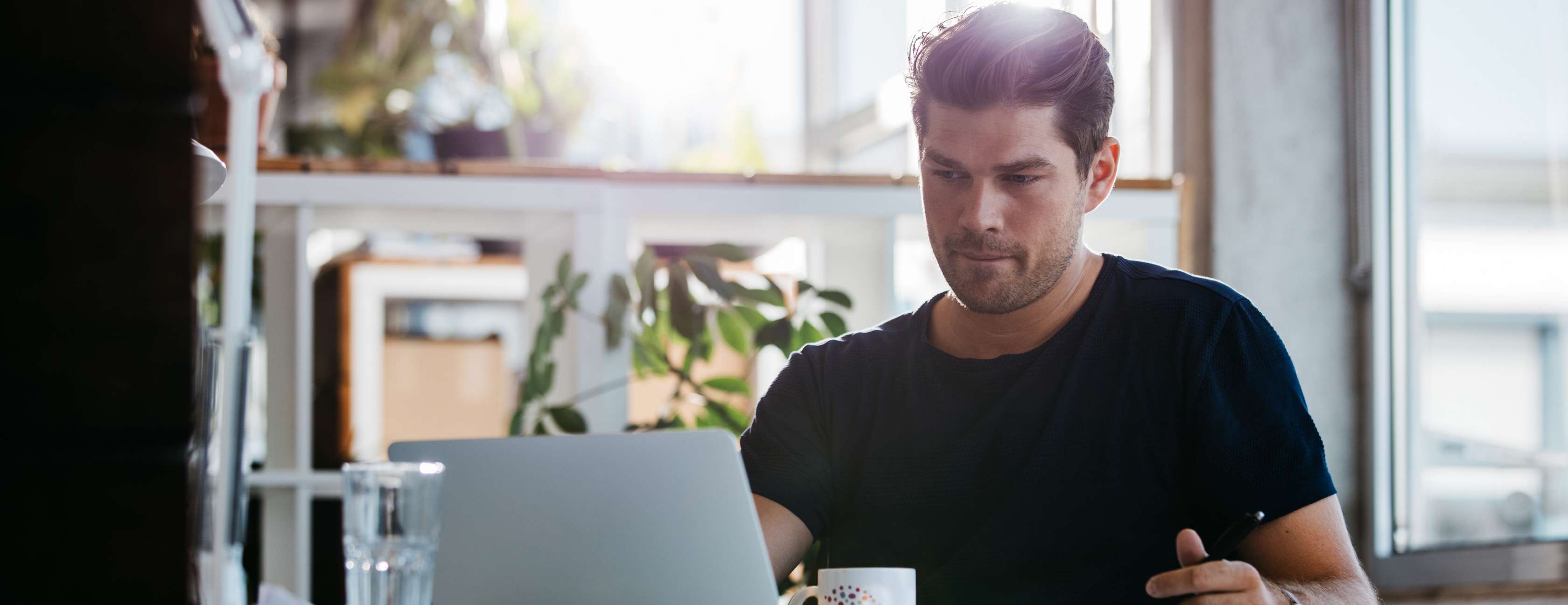 Mann im schwarzen T-Shirt schaut nachdenklich in einen Laptop, vor ihm eine bunte Kaffeetasse
