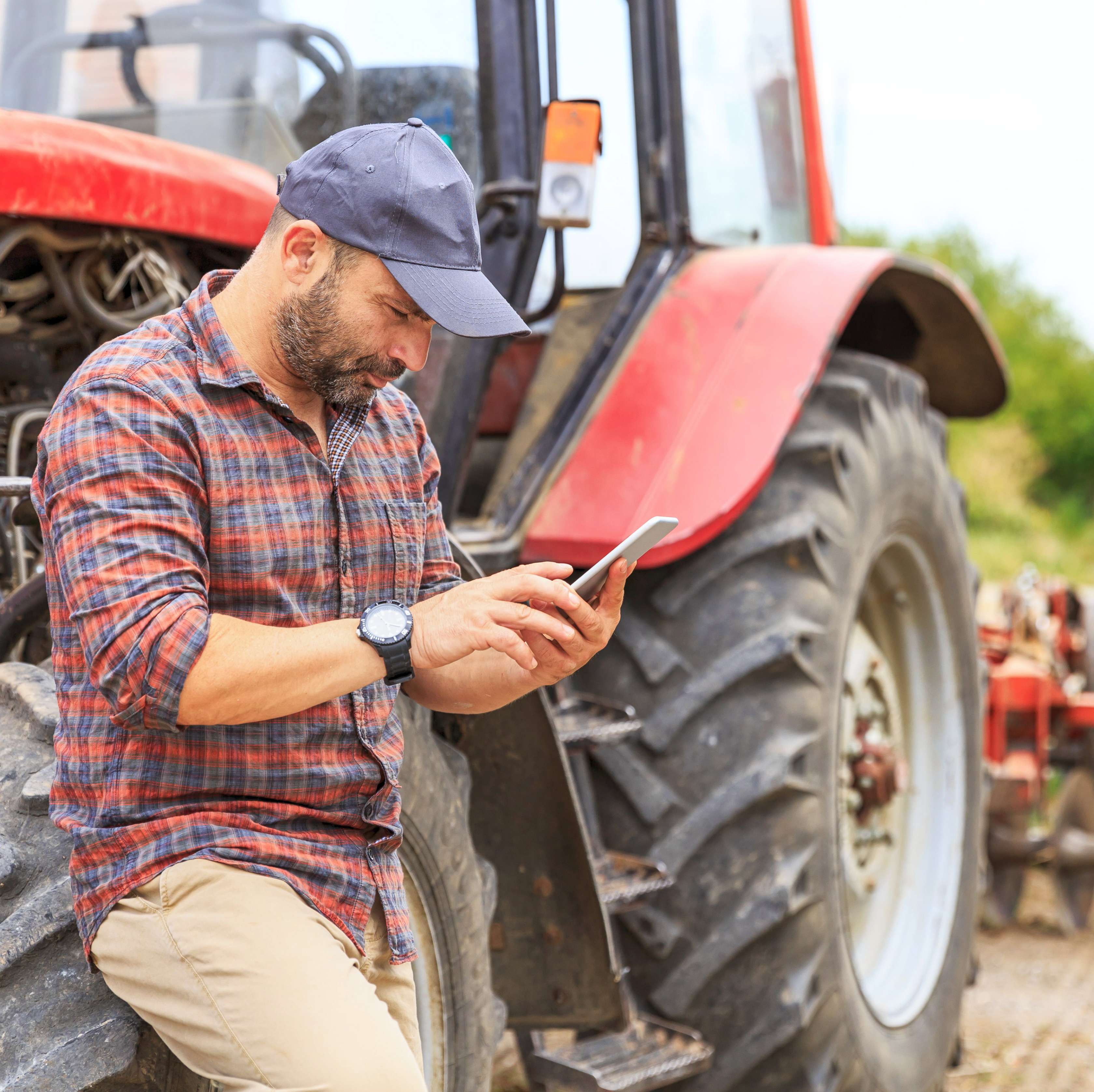Ein Landwirt steht neben einem roten Traktor und schaut in ein Smartphone.