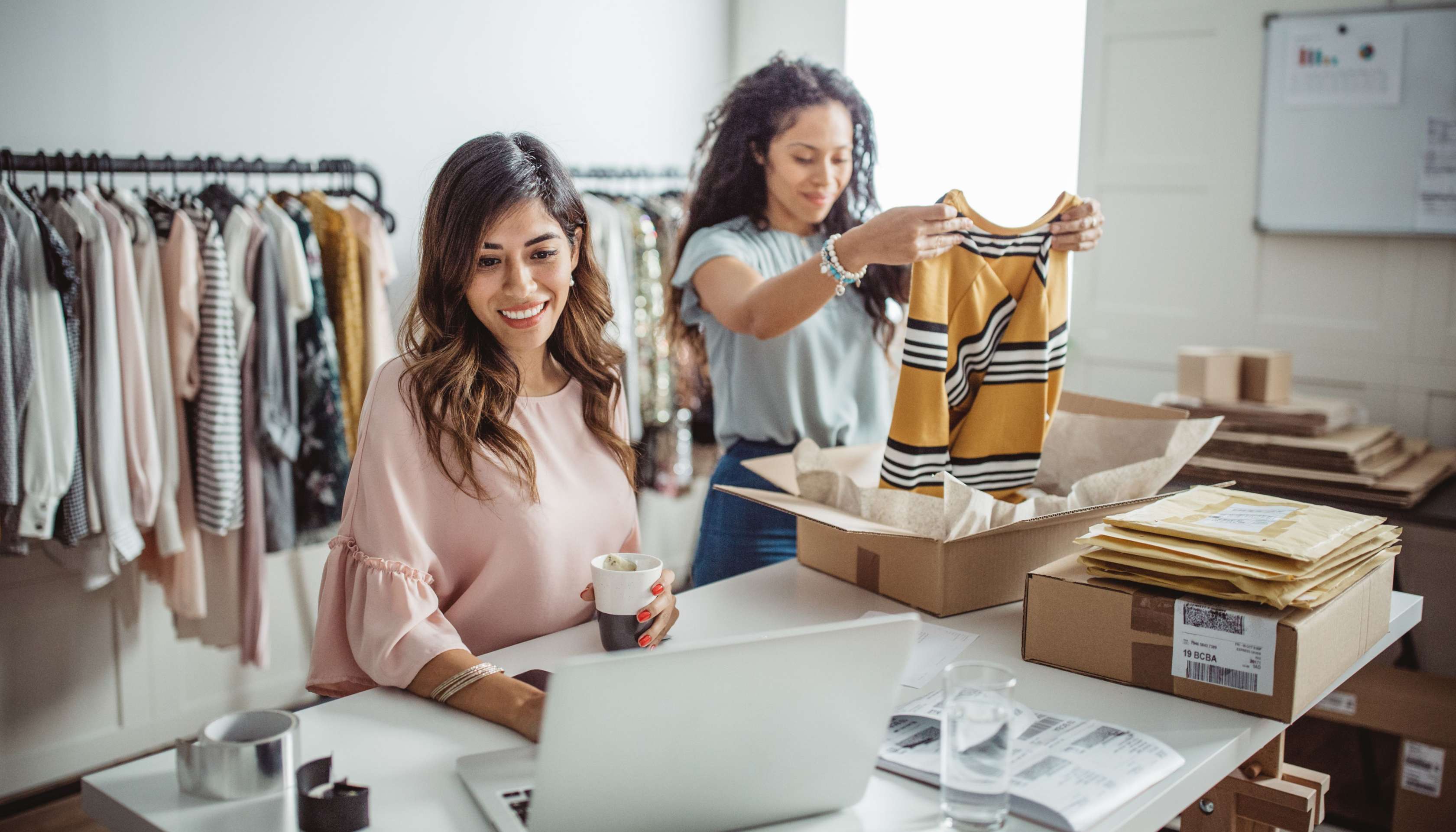 Allianz - Onlineshop gründen | Zwei junge Frauen managen offenbar einen Online-Shop für Kleidung. Eine von ihnen sitzt am Laptop, die andere verpackt Kleidung in einem Karton.