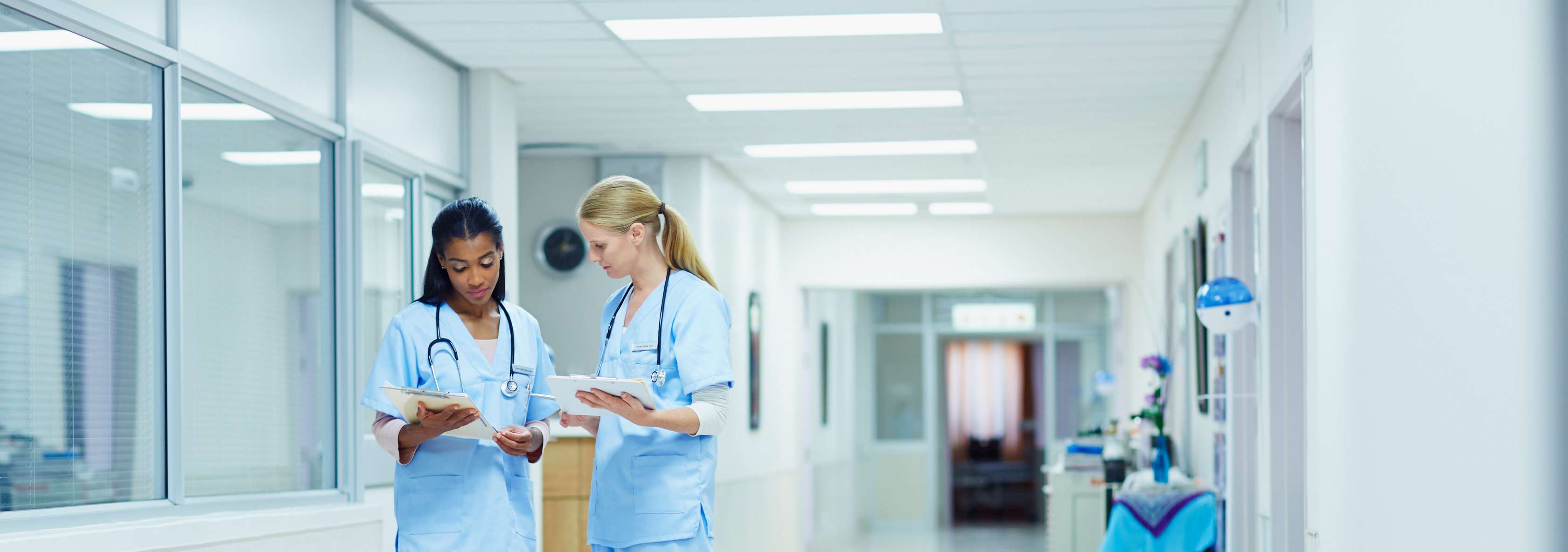 Man sieht zwei Pflegerinnen auf einem Krankenhausflur, die gemeinsam auf ein Klemmbrett schauen.