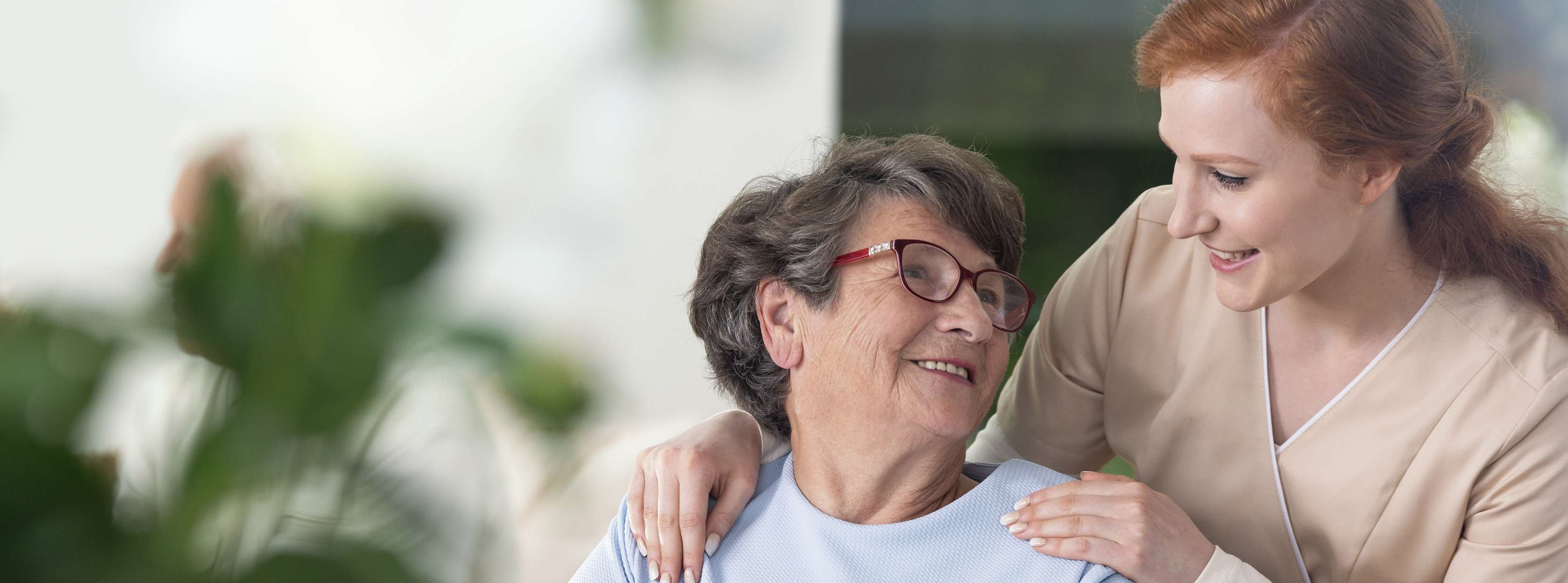 Rothaarige Pflegerin legt lächelnd die Hände auf den Schultern einer älteren Dame