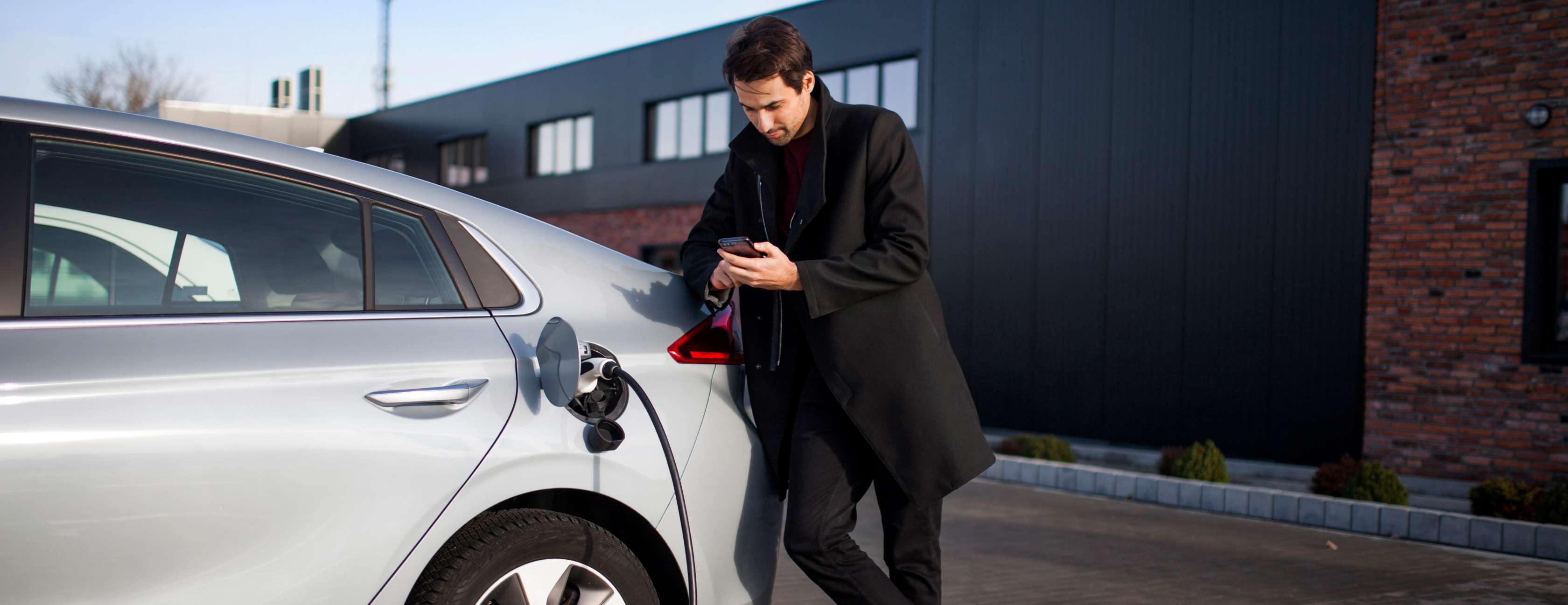 e-Mobility Angebote: Mann steht an einem E-Auto, das gerade aufgeladen wird und schaut in sein Mobiltelefon.