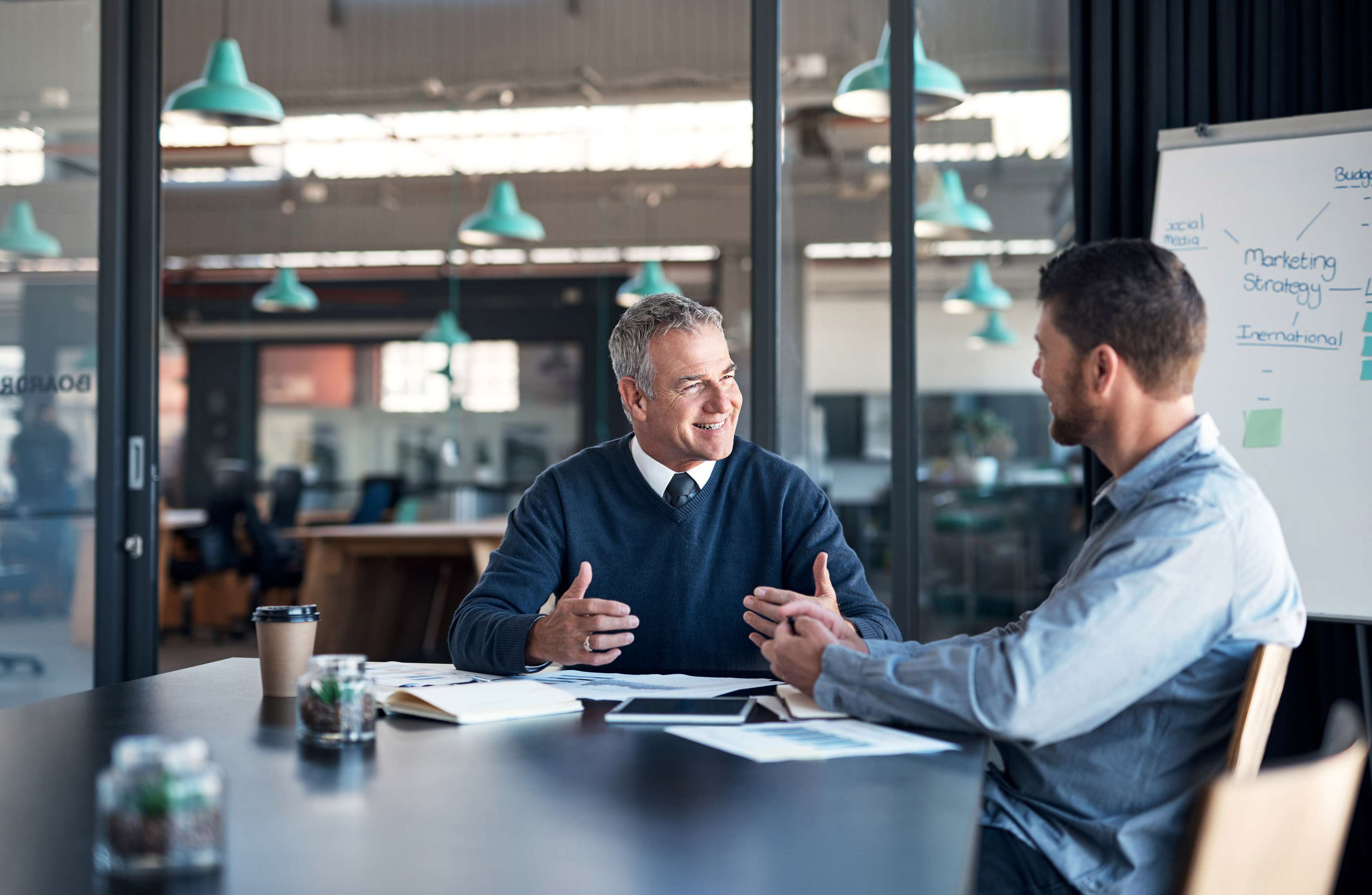 Zwei Männer sitzen am Konferenztisch und sprechen miteinander, hinter ihnen ein Chart mit einer Marketingstrategie