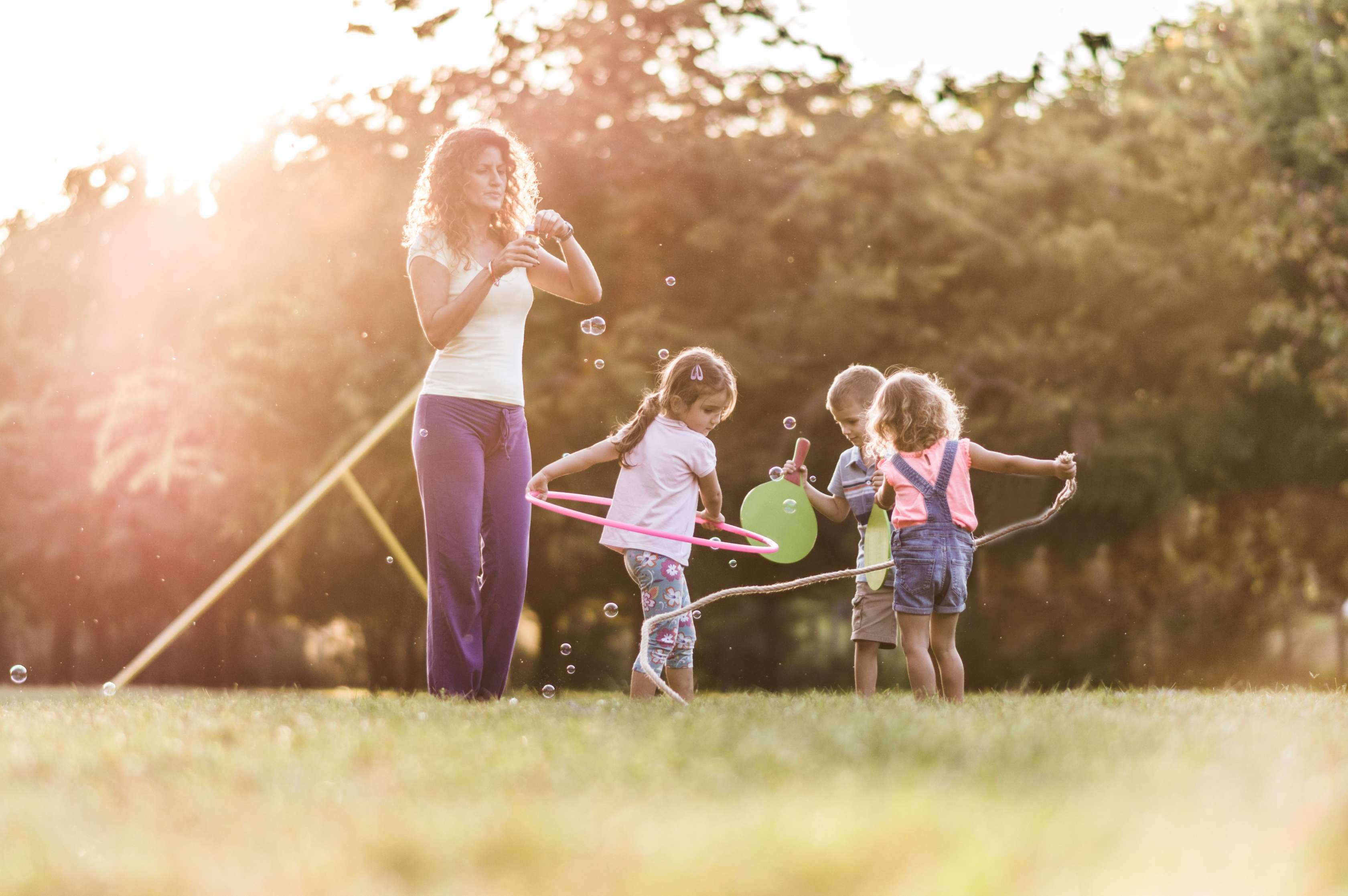 Eine junge Kindergärtnerin steht mit drei spielenden Kindern im Garten und bläst Seifenblasen.