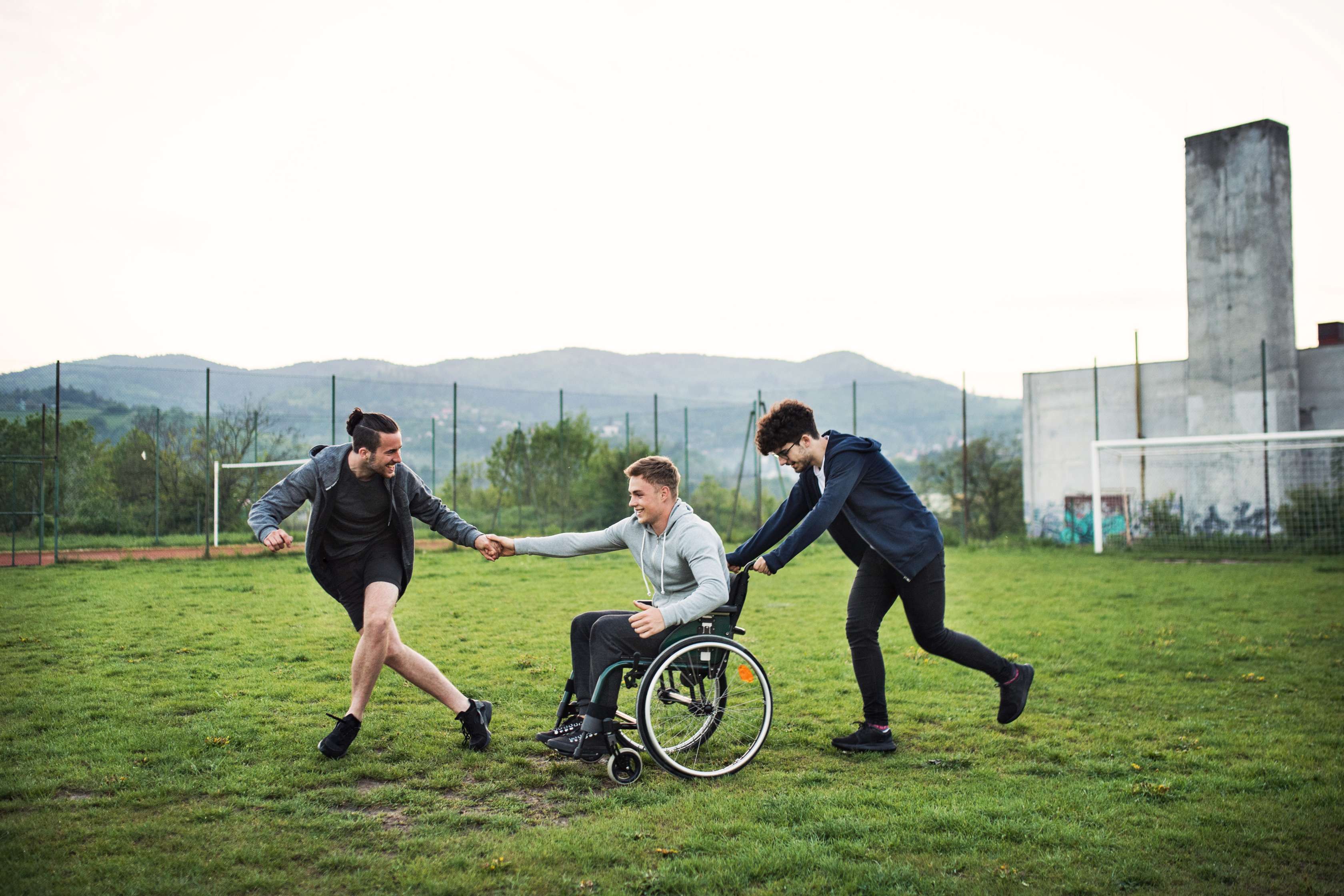 Junger Mann im Rollstuhl wird lachend von seinen Freunden auf einem Fussballfeld gezogen und geschoben.