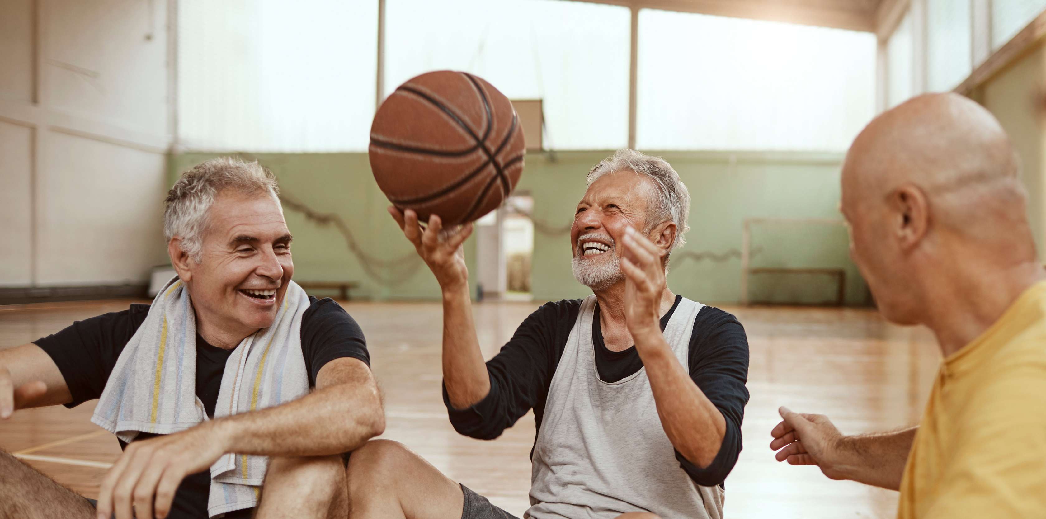 Drei ältere Männer sitzend lachend in Sportkleidung auf dem Boden, während einer von ihnen einen Basketball hochhält