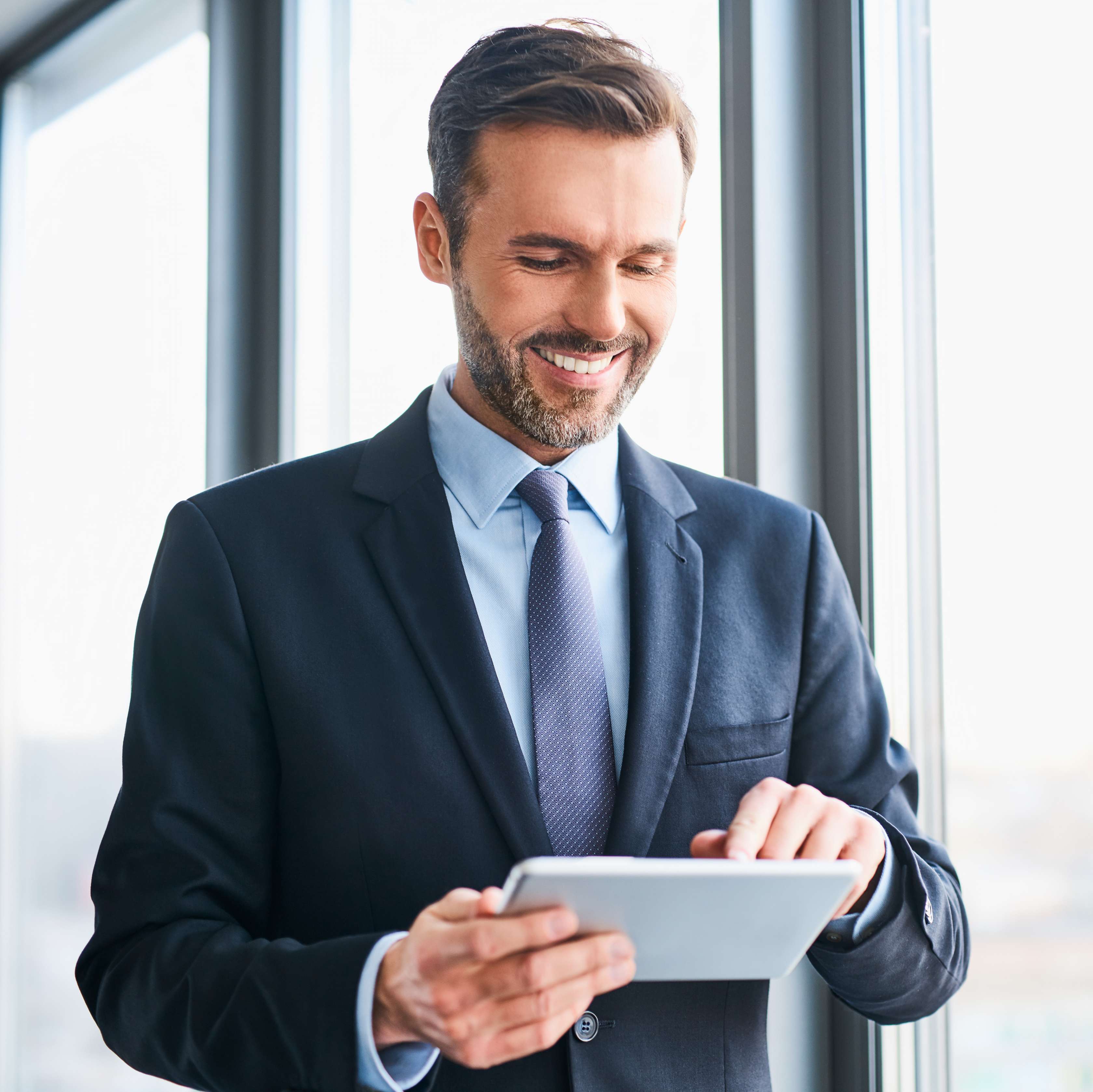 Ein Geschäftsmann mit Bart und blauem Anzug schaut lächelnd auf ein Tablet, das er in der Hand hält