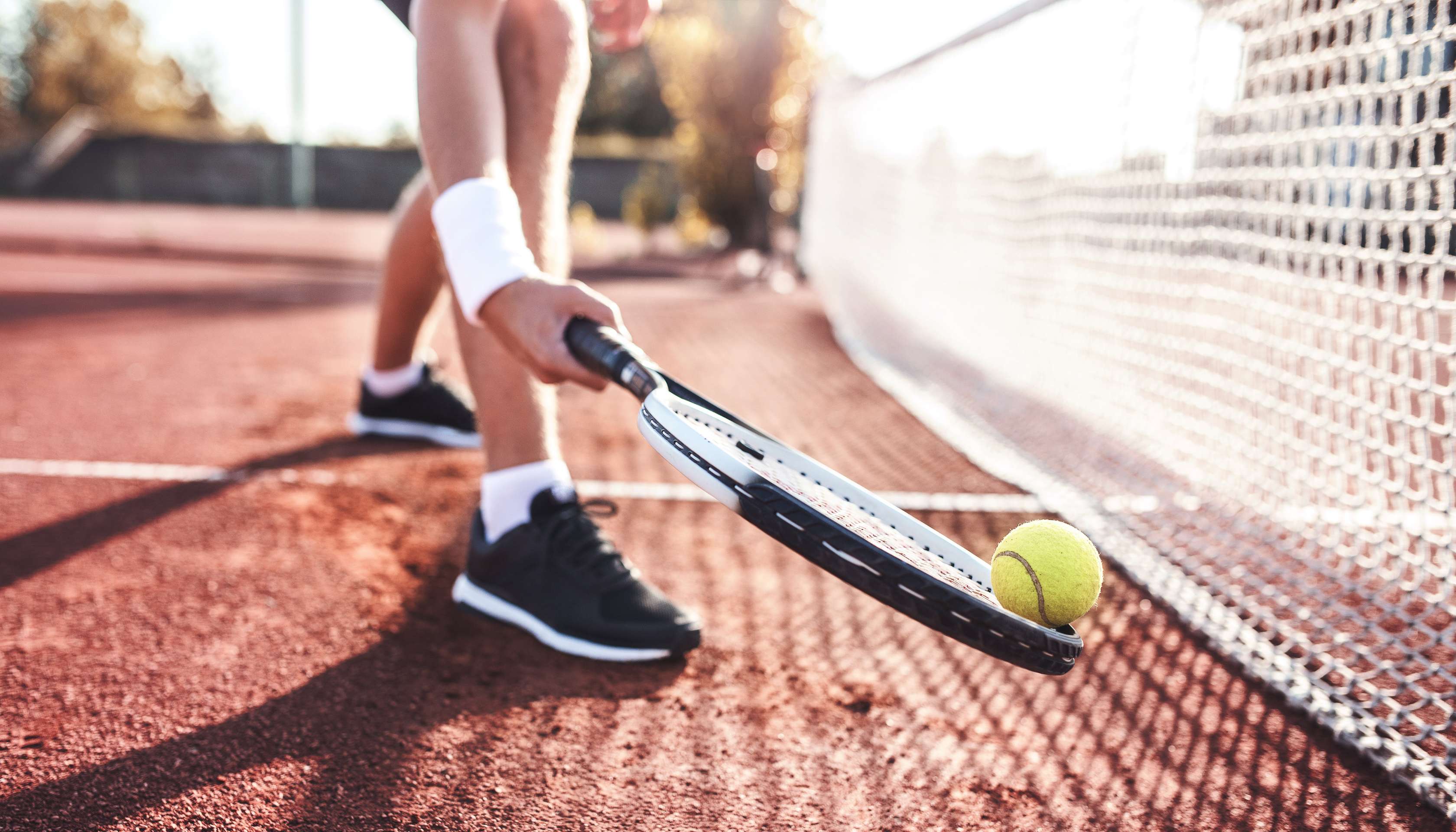 Spieler hebt Tennisball mit Tennisschläger vor dem Netz auf, nur die Füße sind zu sehen.