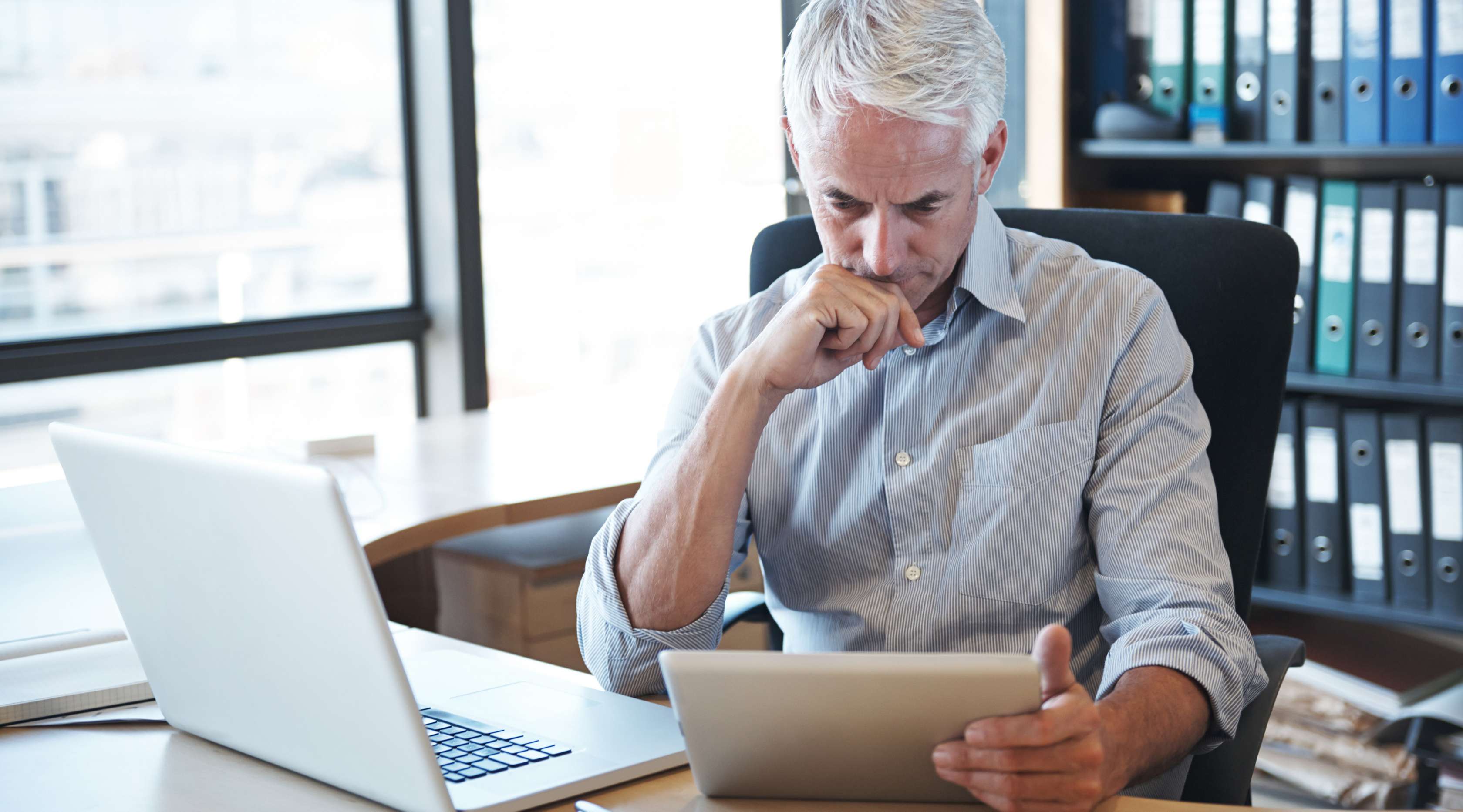 Ein Mann sitzt neben einem aufgeklappten Laptop an einem Schreibtisch in einem Büro und blickt nachdenklich in sein Tablet, welches er in seiner Hand hält.