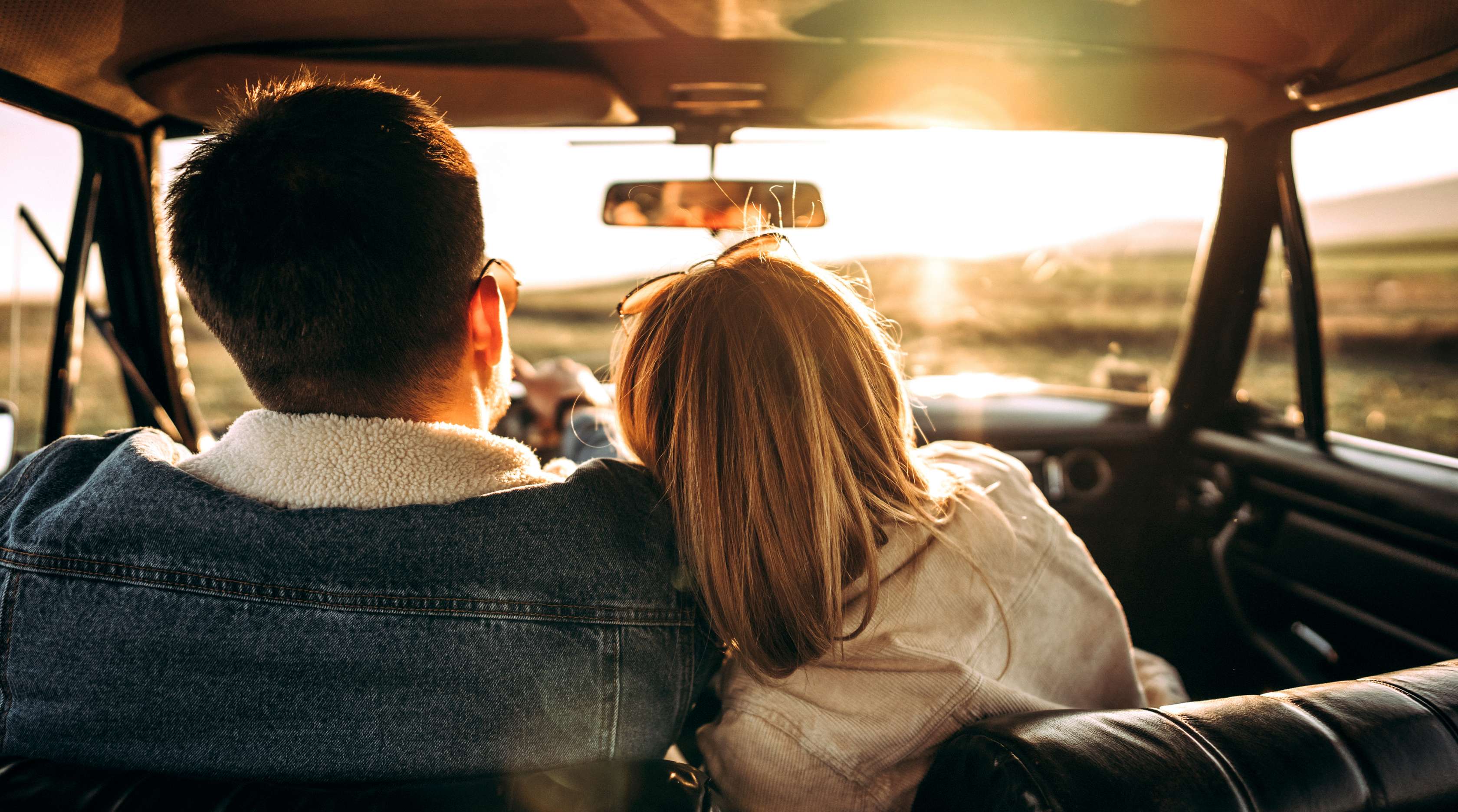 Allianz - Fahrerschutzversicherung: Paar sitzt aneinander gelehnt in Auto und blickt in den Sonnenuntergang