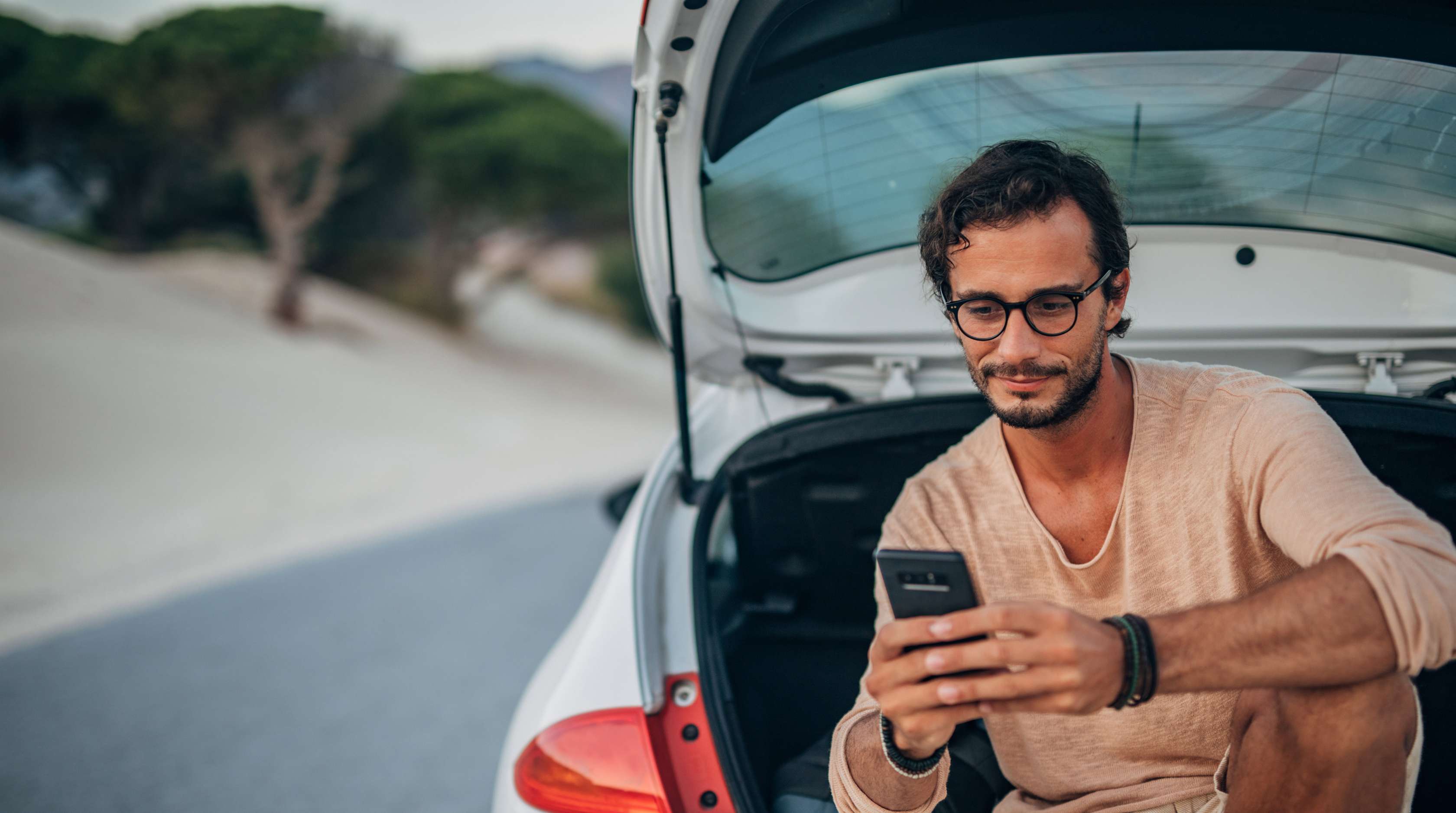 Mann mit Brille und Bart sitzt im Kofferraum seines Autos und schaut ins Smartphone