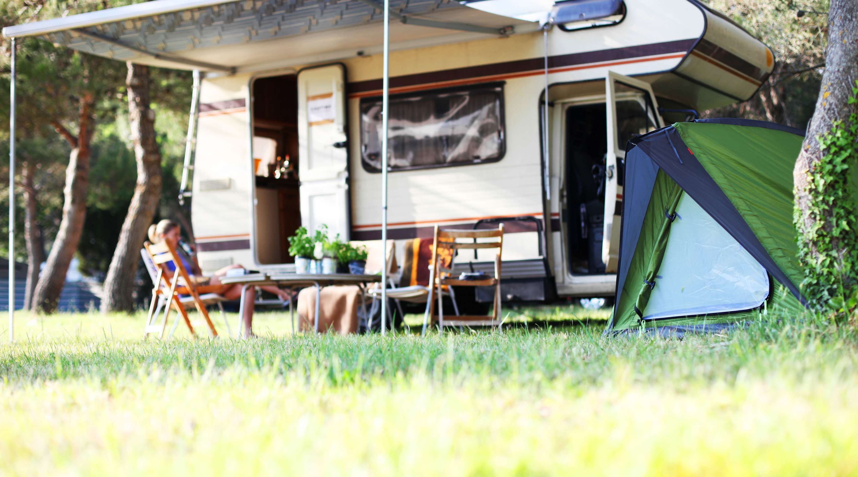 Wohnmobil mit ausgefahrener Markise und Camping-Möbeln steht auf Wiese neben Zelt