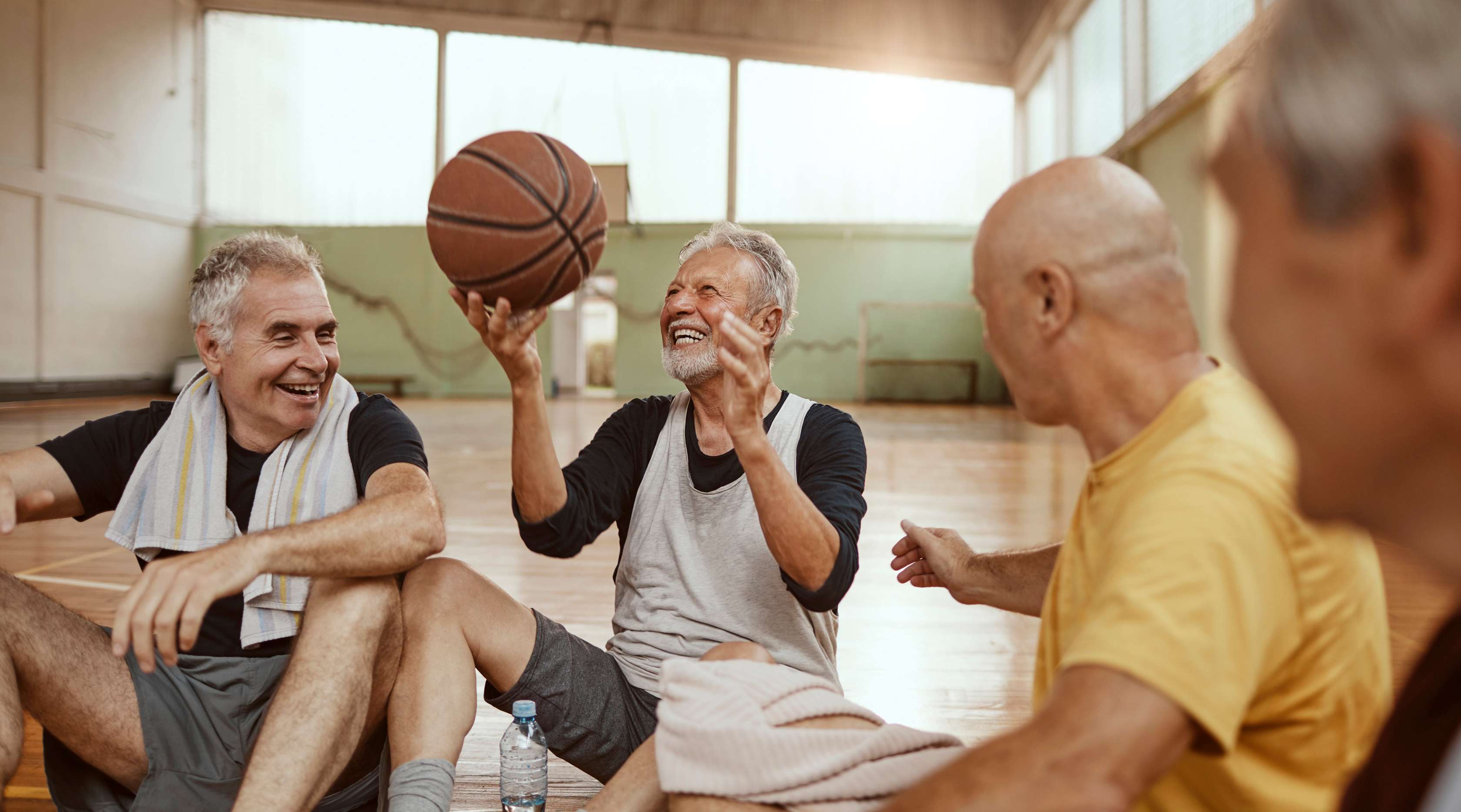 Ältere Männer sitzen im Kreis auf dem Boden einer Turnhalle und lachen, einer von ihnen hält einen Basketball hoch.