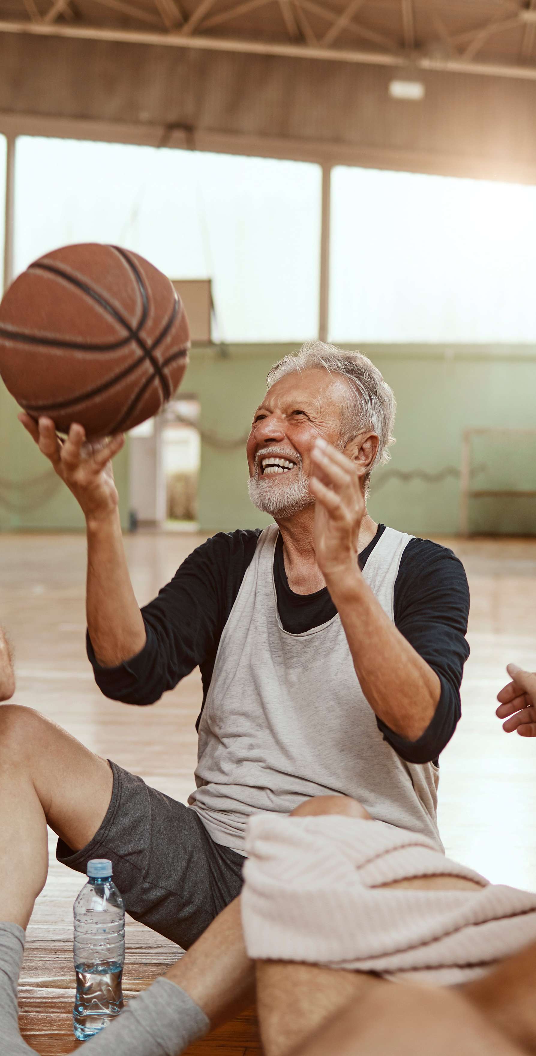 Ältere Männer sitzen im Kreis auf dem Boden einer Turnhalle und lachen, einer von ihnen hält einen Basketball hoch.