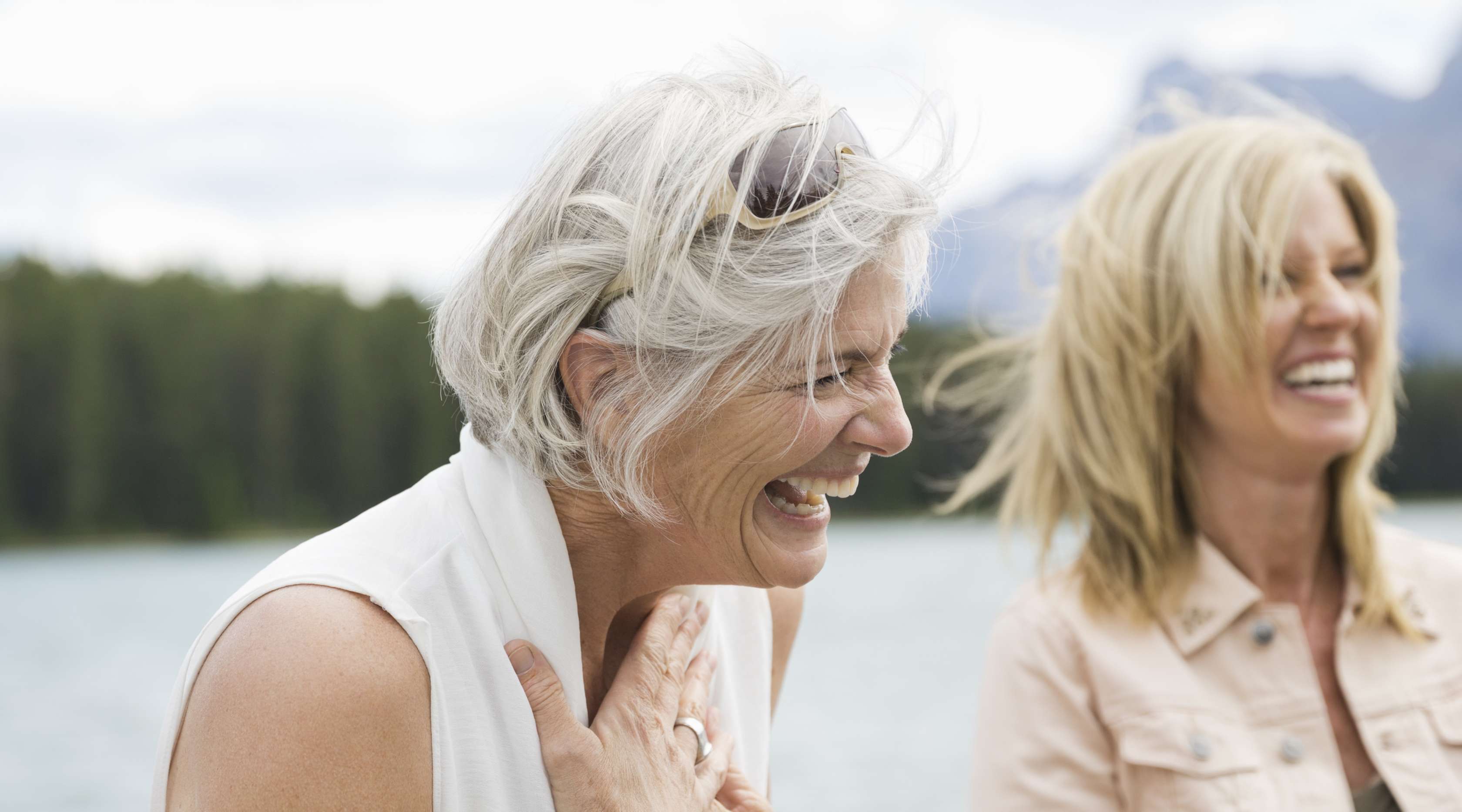 Seniorin und Frau mittleren Alters stehen vor einem See und lachen 