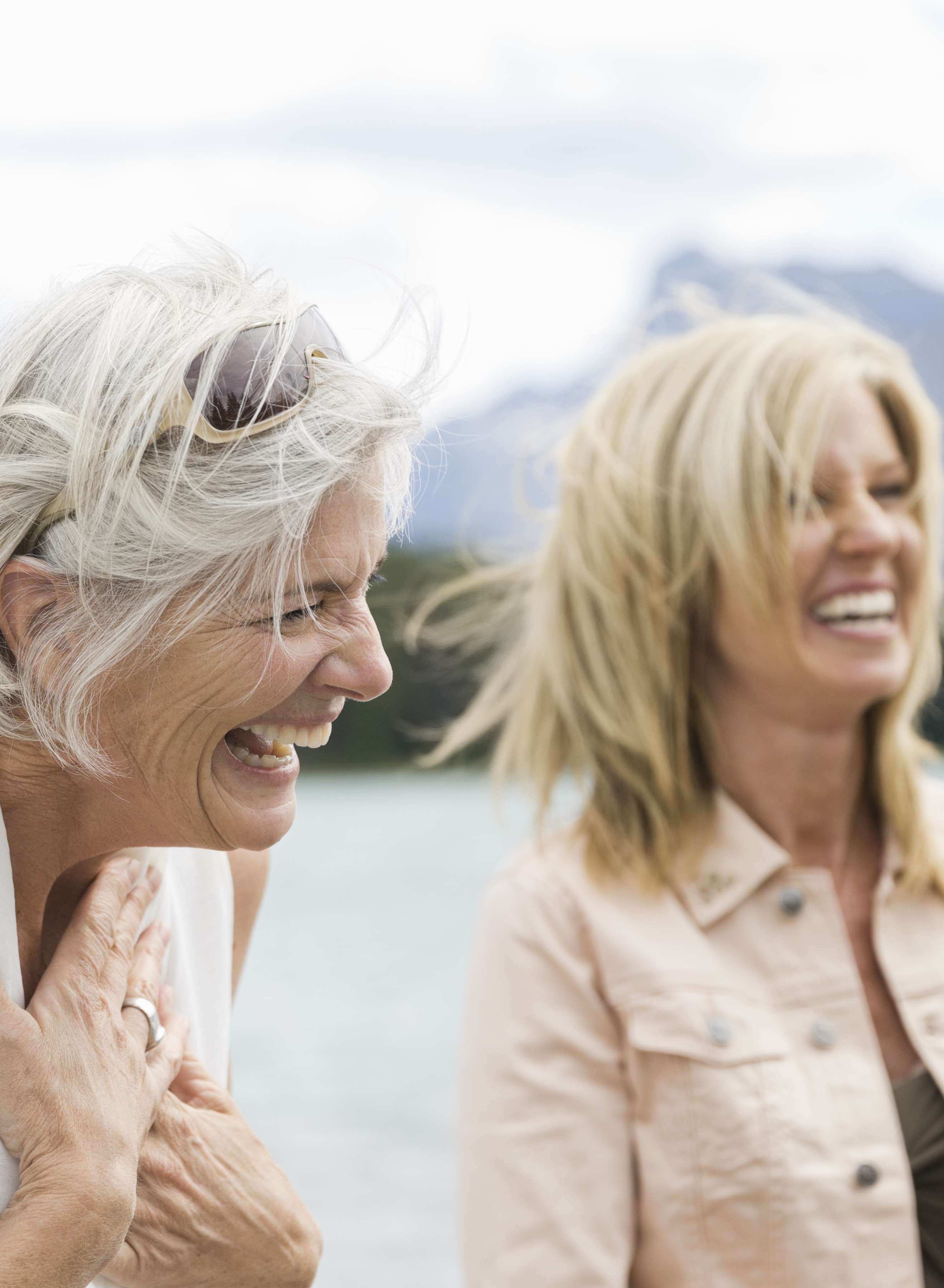 Seniorin und Frau mittleren Alters stehen vor einem See und lachen 