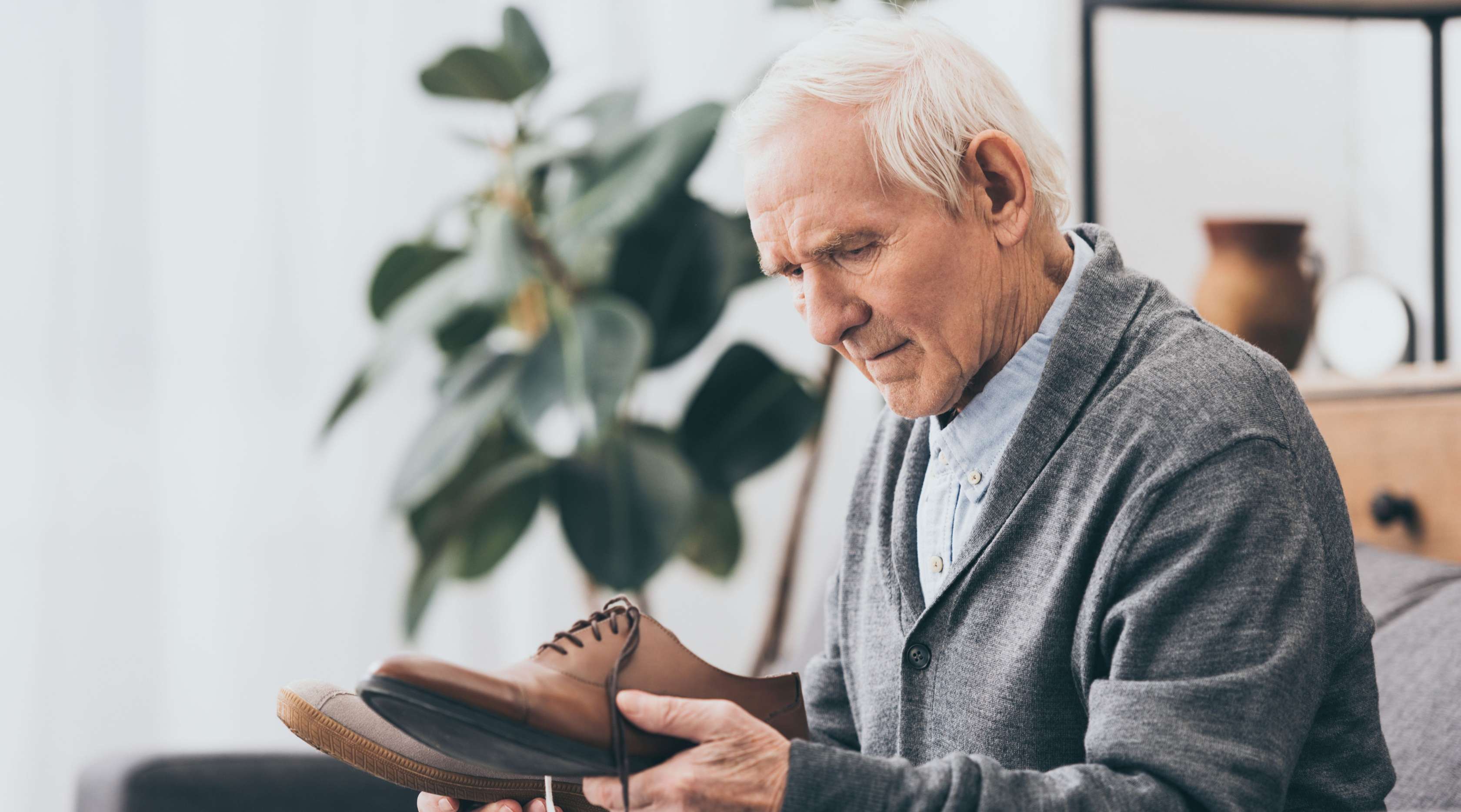 Älterer Mann mit weißem Haar sitzt ratlos mit einem Paar brauner Schuhe in der Hand auf dem Sofa.