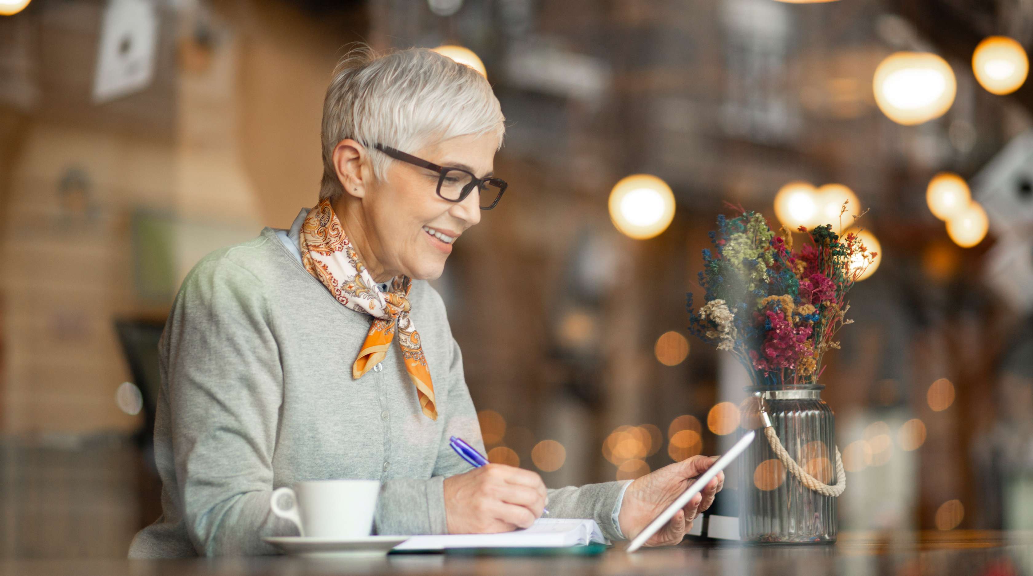 Ältere Frau sitzt lächelnd in einem Cafe und macht Notizen