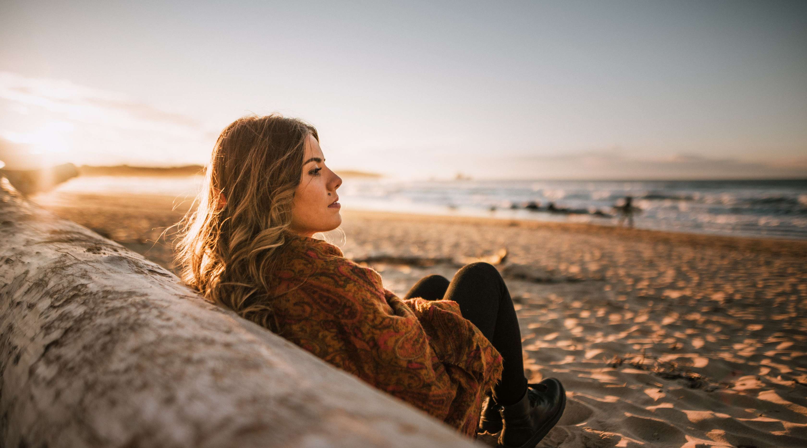 Frau sitzt am Strand angelehnt an einem Baumstamm und schaut aufs Meer hinaus
