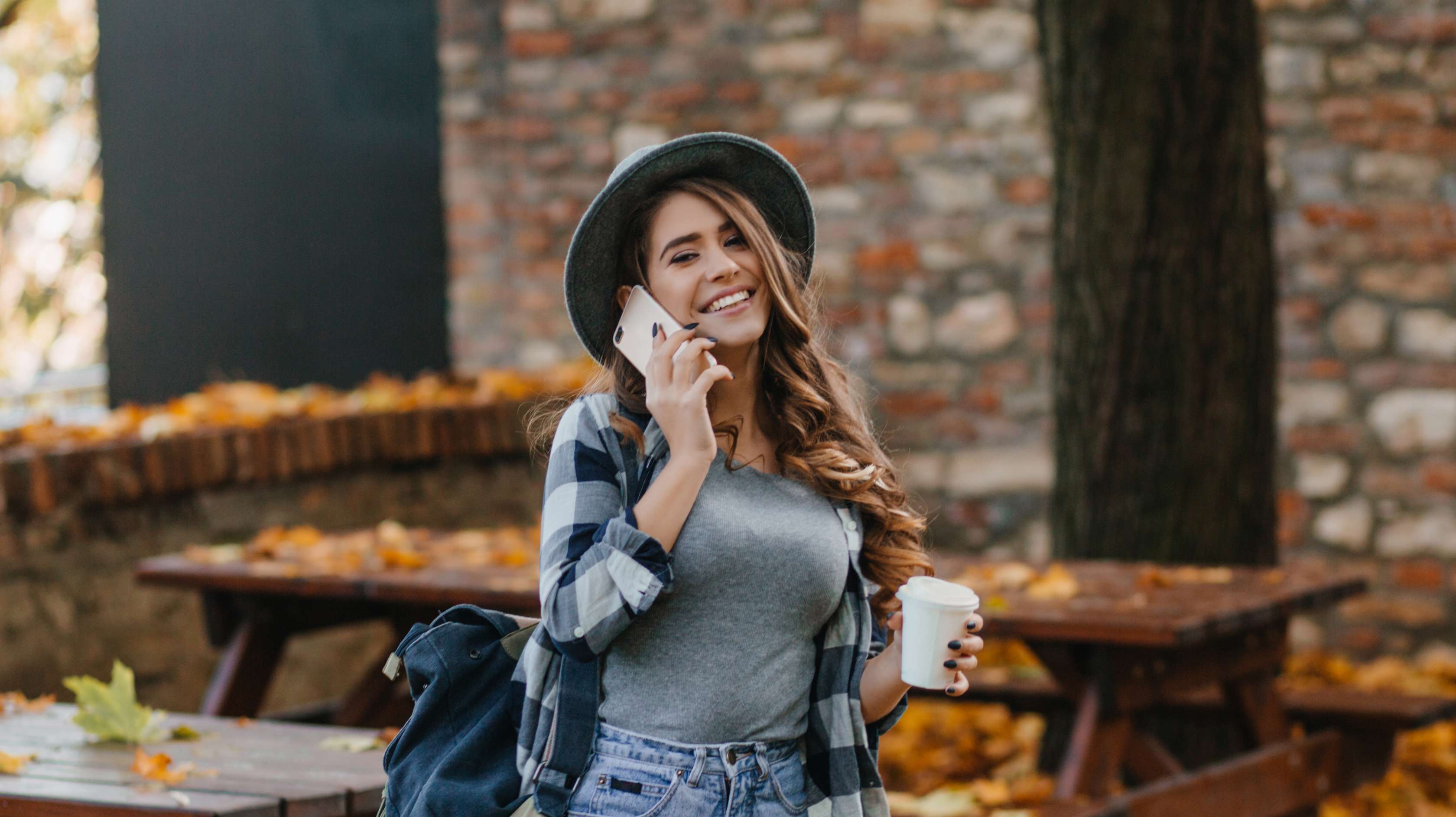 Junge Frau steht draußen auf Laubboden mit Holztischen im Hintergrund und lächelt beim Telefonieren