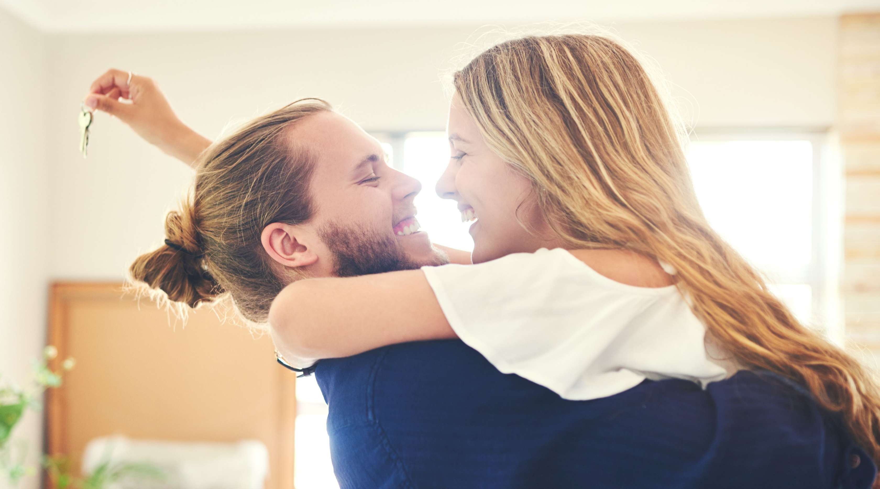 Ein junges Paar hält sich im Arm und lächelt sich fröhlich an. Im Hintergrund sieht man ihr neues Haus, sie hält den neuen Hausschlüssel in der Hand.