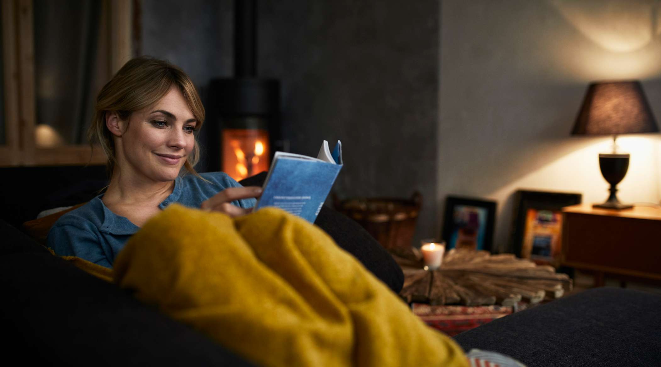 Allianz - Hausratversicherung - Sengschaden: Frau liegt mit Buch auf ihrer Couch im Wohnzimmer vorm Kamin