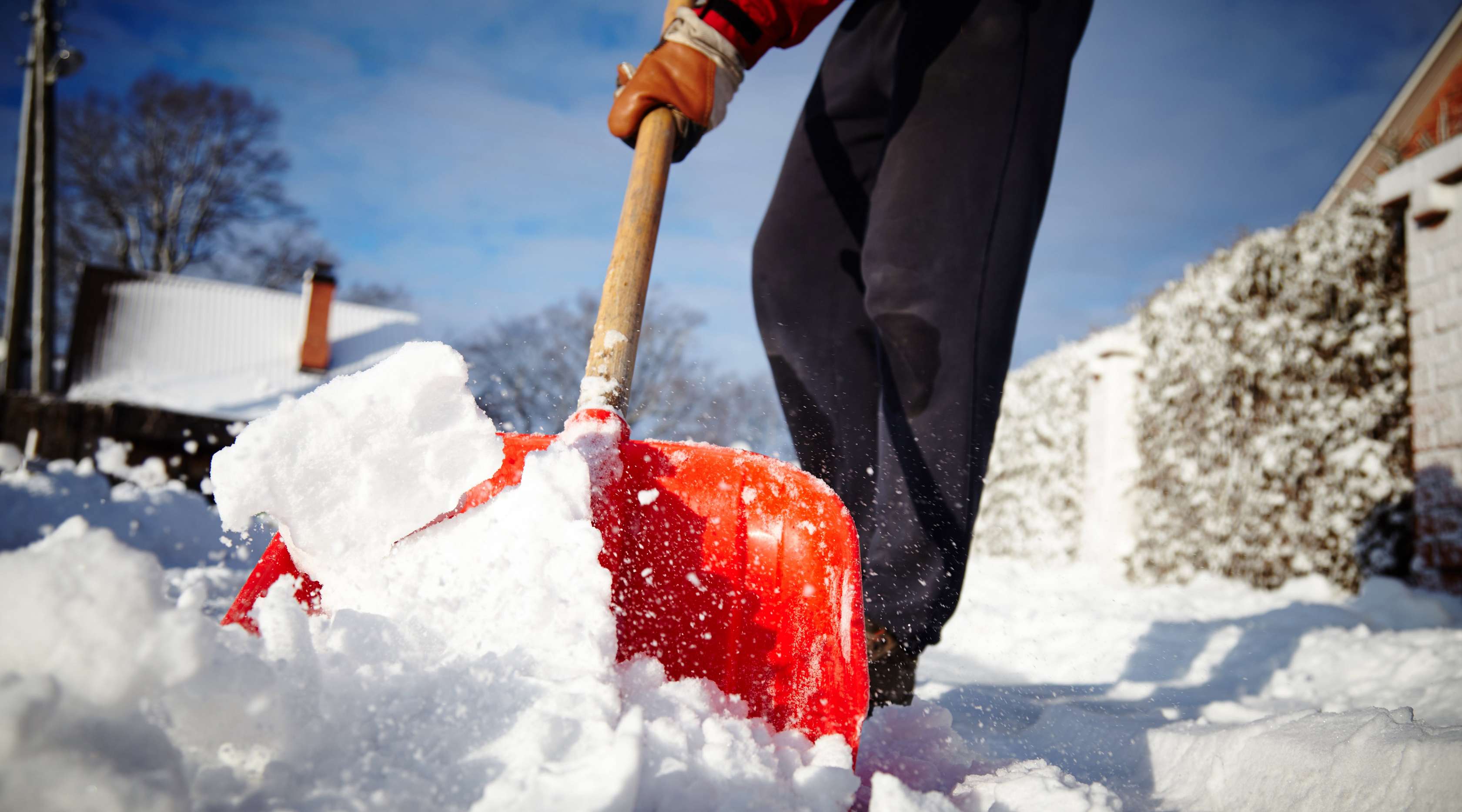 Mann in schwarzer Hose räumt mit roter Schneeschippe den scheebedeckten Gehweg vor Hauseingang frei