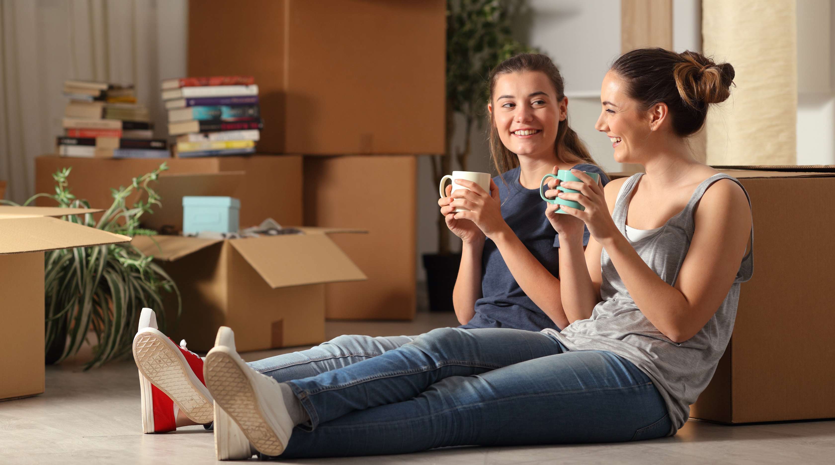 Zwei junge Frauen sitzen auf dem Boden trinken einen Kaffee und lachen im Hintergrund Umzugskartons