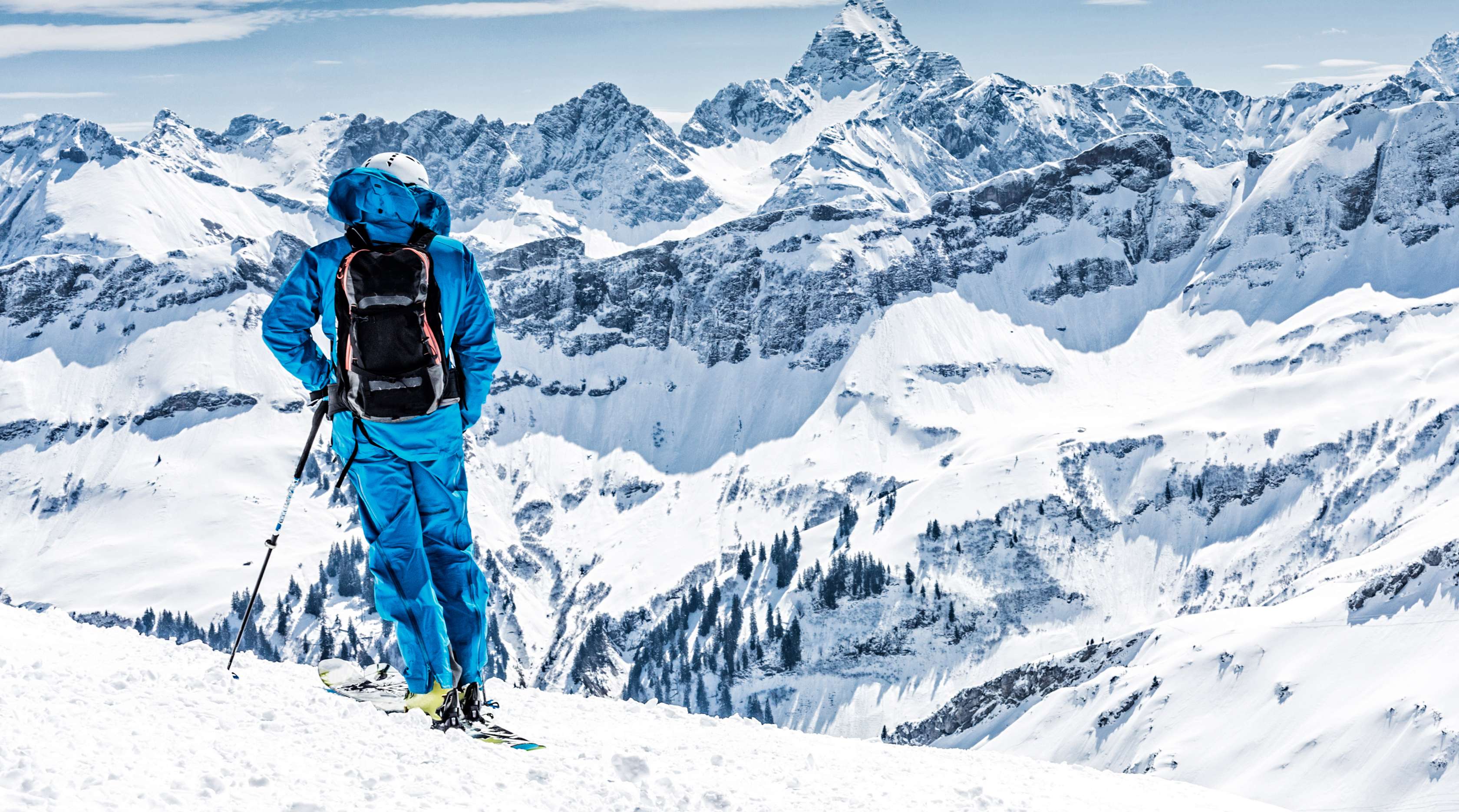 Skifahrer in blauem Skianzug steht auf der Piste und blickt auf das verschneite Gebirge.