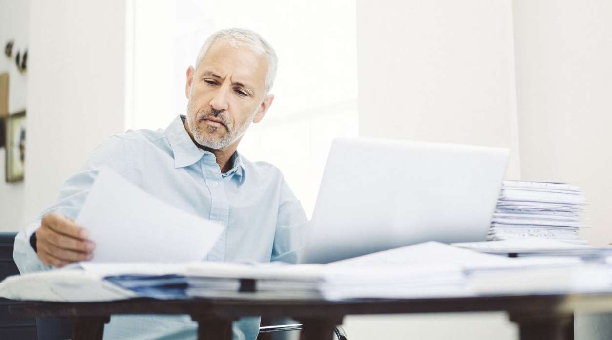 Mann sitzt mit einem Dokument in der Hand vor einem Schreibtisch, auf dem ein Laptop steht