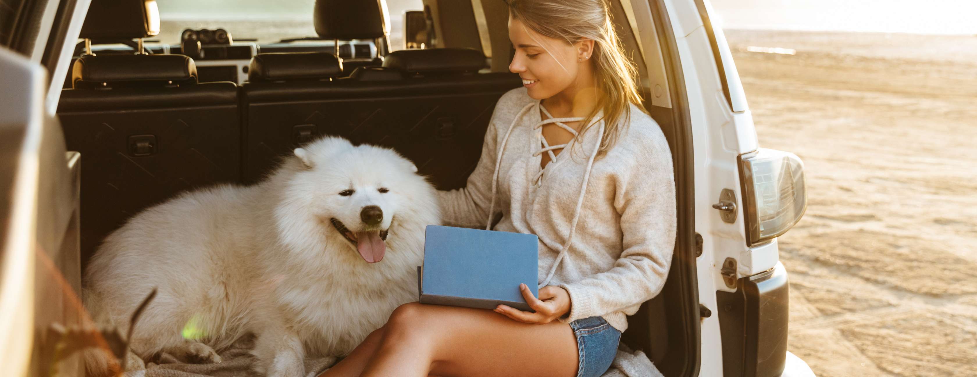Frau sitzt mit einem Hund im Kofferraum ihres SUVs, der am Strand steht