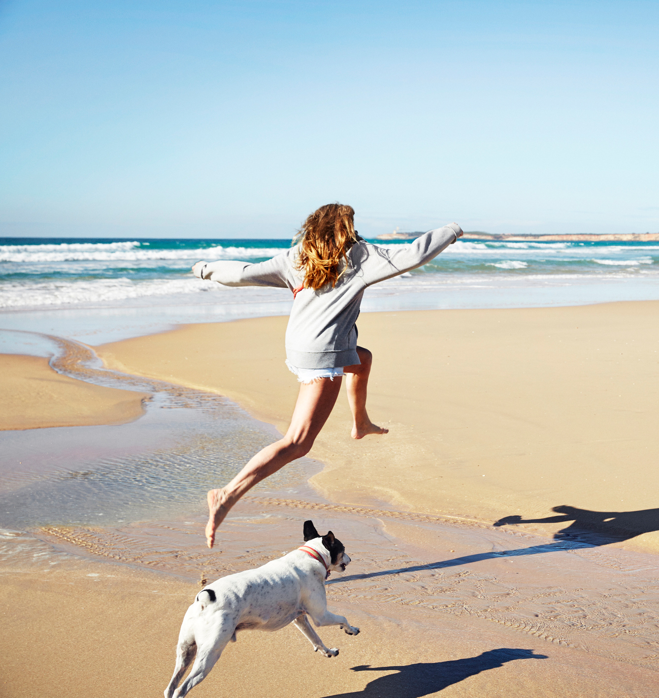 Allianz - Urlaub mit Hund: Frau springt mit Hund über den Strand