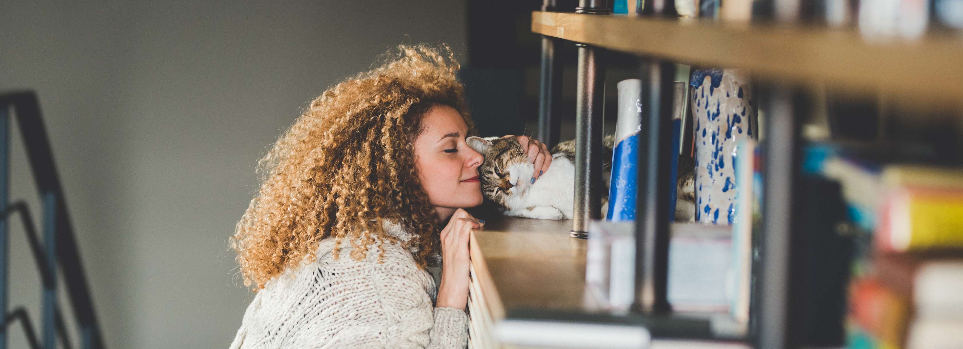 Allianz Katzenversicherung – Welche Katze passt zu mir: Frau mit Locken schmust mit einer auf einem Regal liegenden Katze