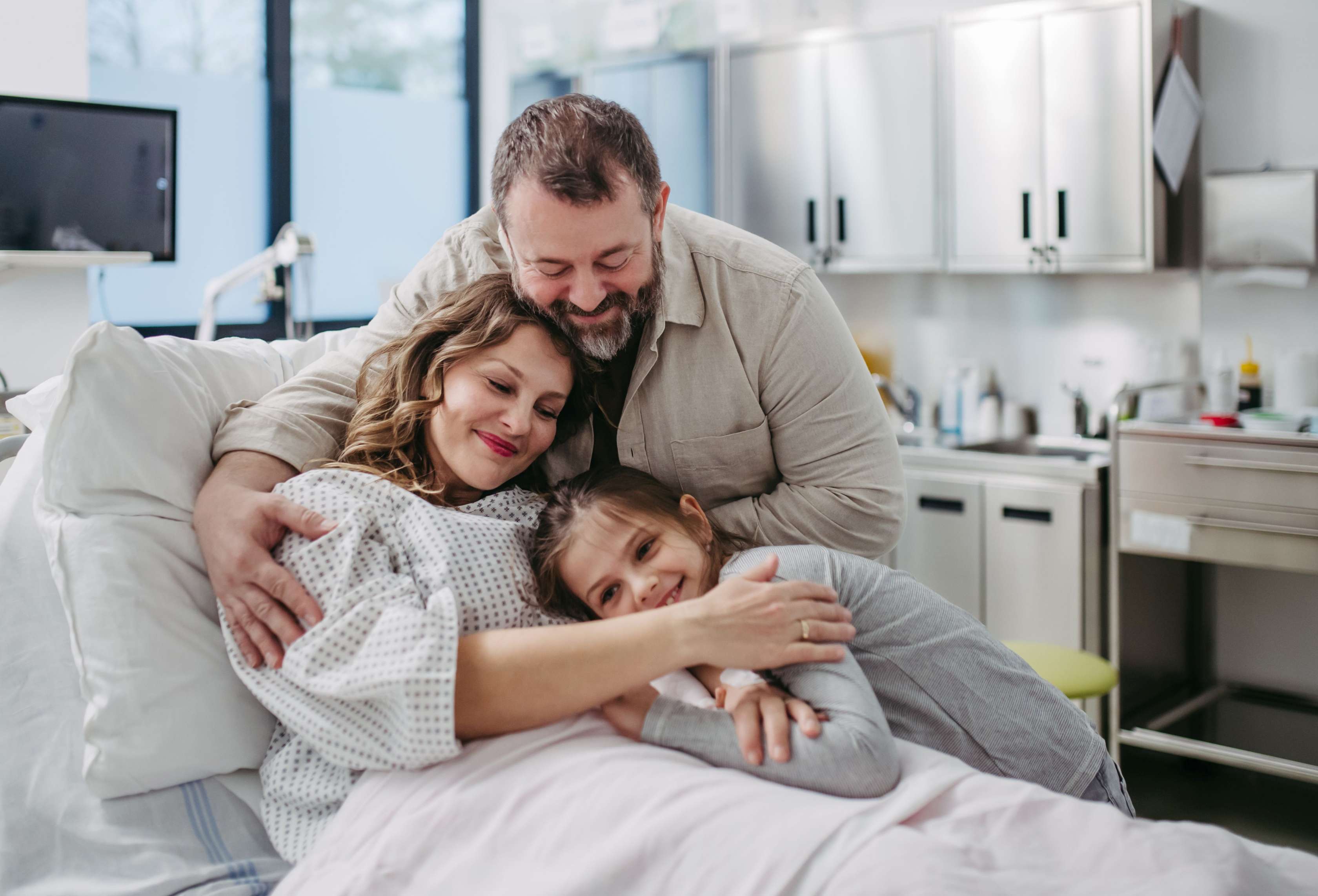 Allianz Krankenzusatzversicherung für Familien: Eine Frau im Krankenhausbett wird von ihrem Mann und ihrem Kind umarmt.
