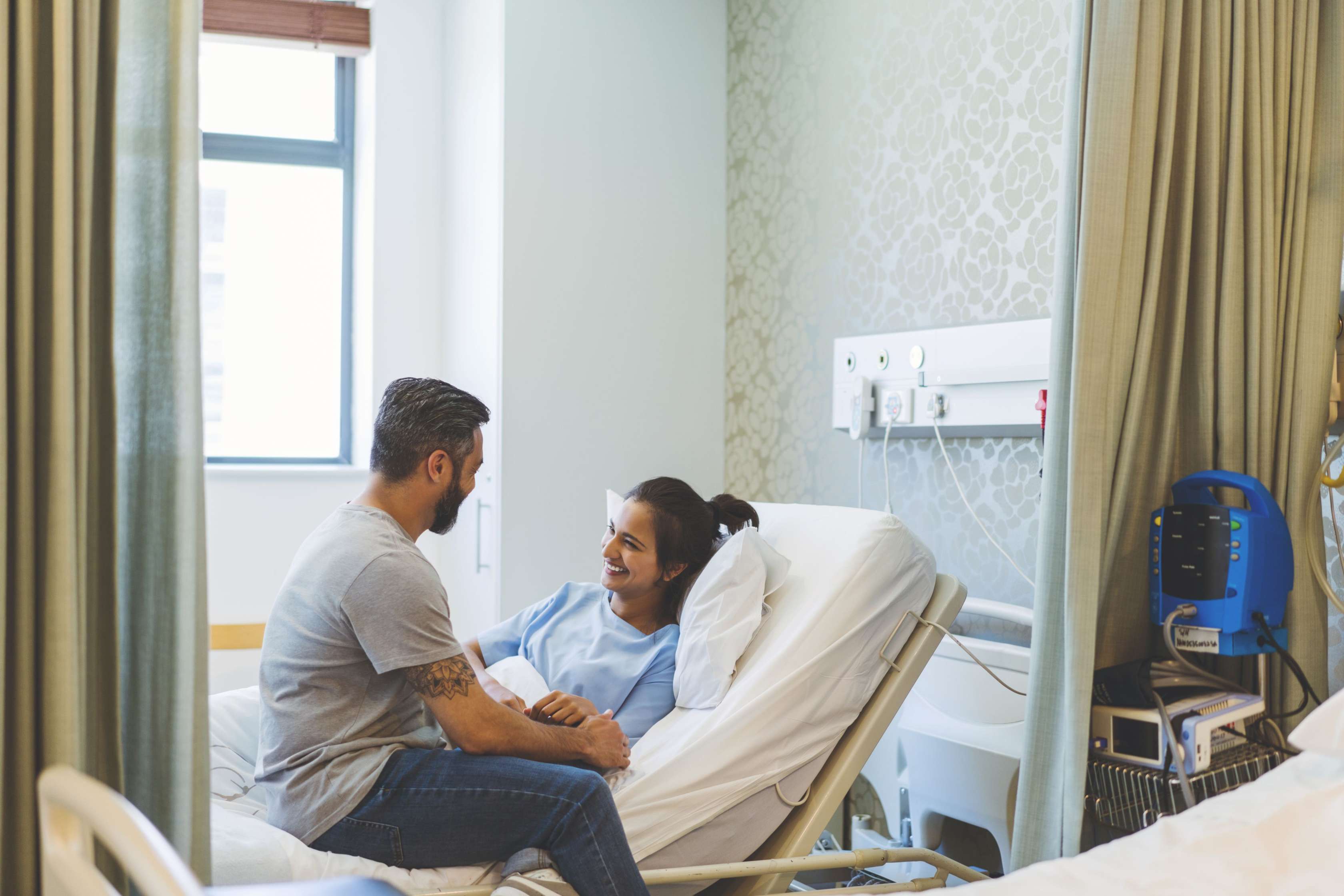 Eine Frau liegt in einem Krankenhausbett, ein Mann sitzt ihr zugewandt auf der Kante des Bettes. Die beiden sehen sich an und lächeln.