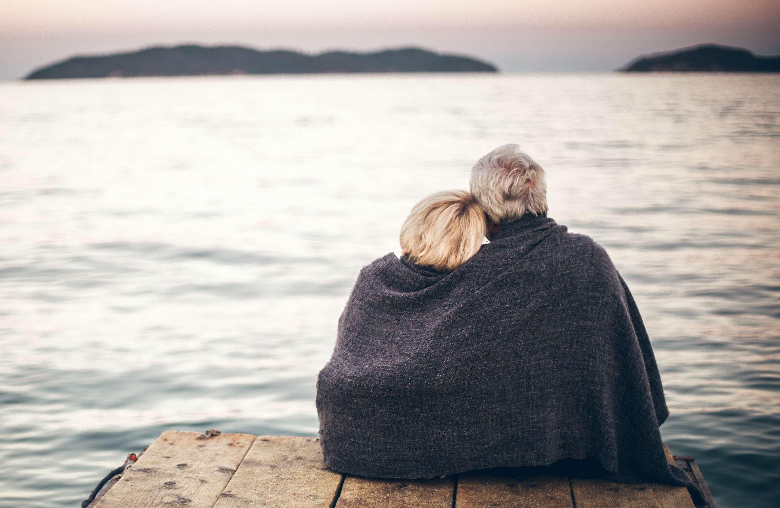 Allianz - Pflegerentenversicherung : Älteres Paar mit grauer Decke über dem Rücken schaut sich das Meer an