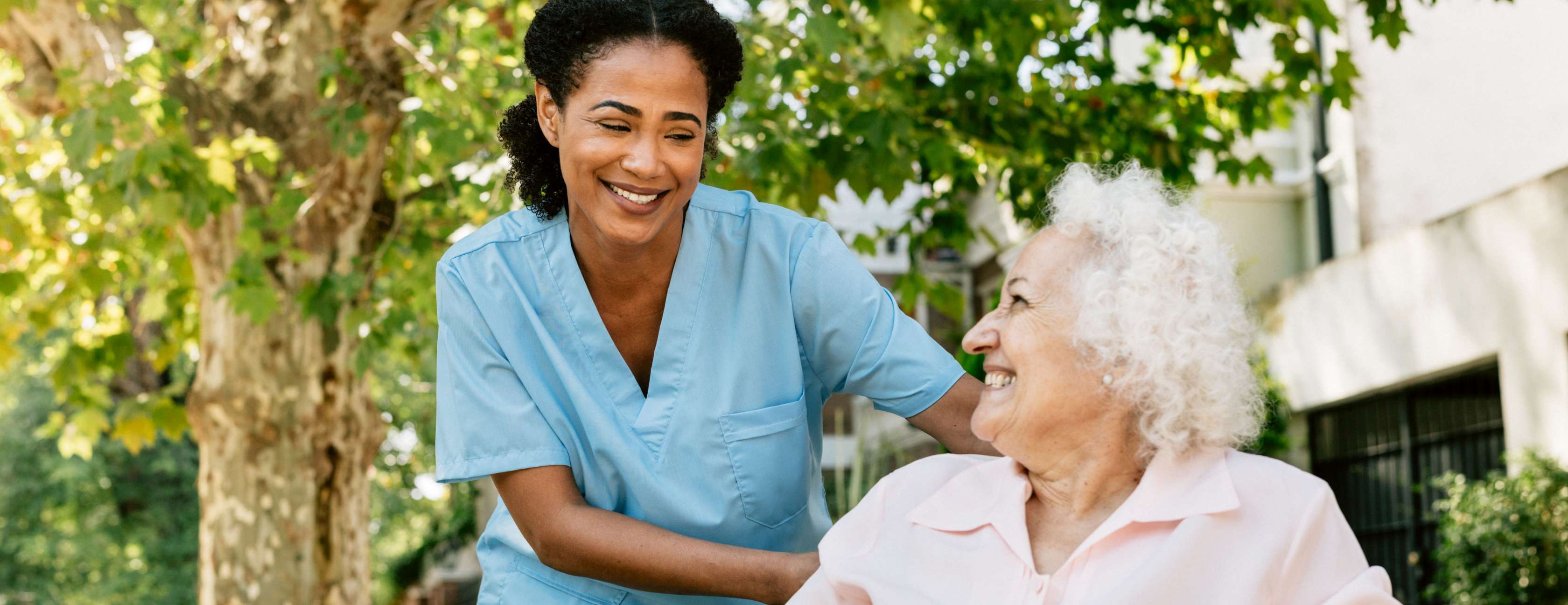Pflegereform 2027: Eine Pflegerin schiebt eine Seniorin im Rollstuhl auf dem Gehweg und lächelt sie an.