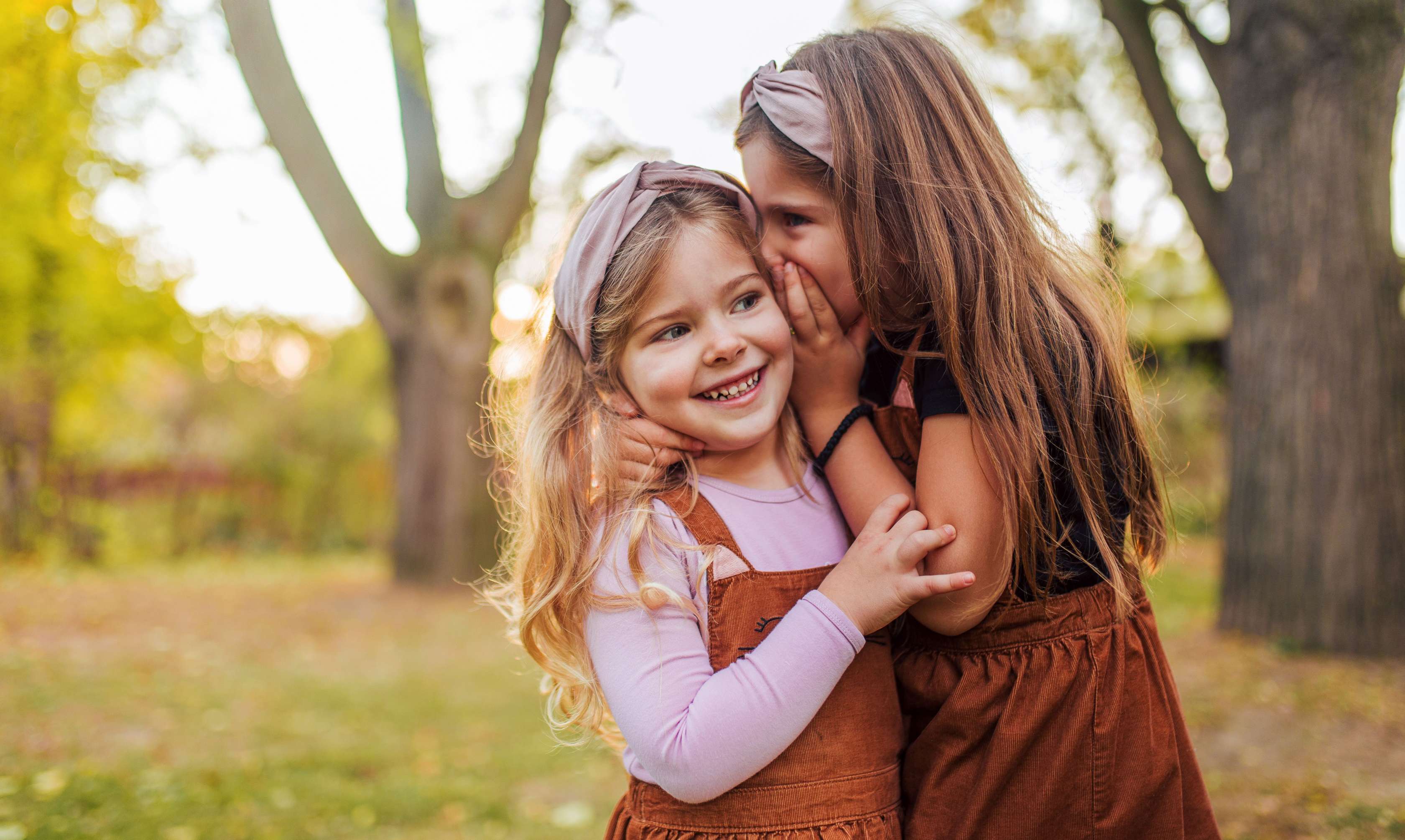 Zwei Mädchen stehen vergnügt in einem herbstlichen Wald. Eine flüstert der anderen ins Ohr.