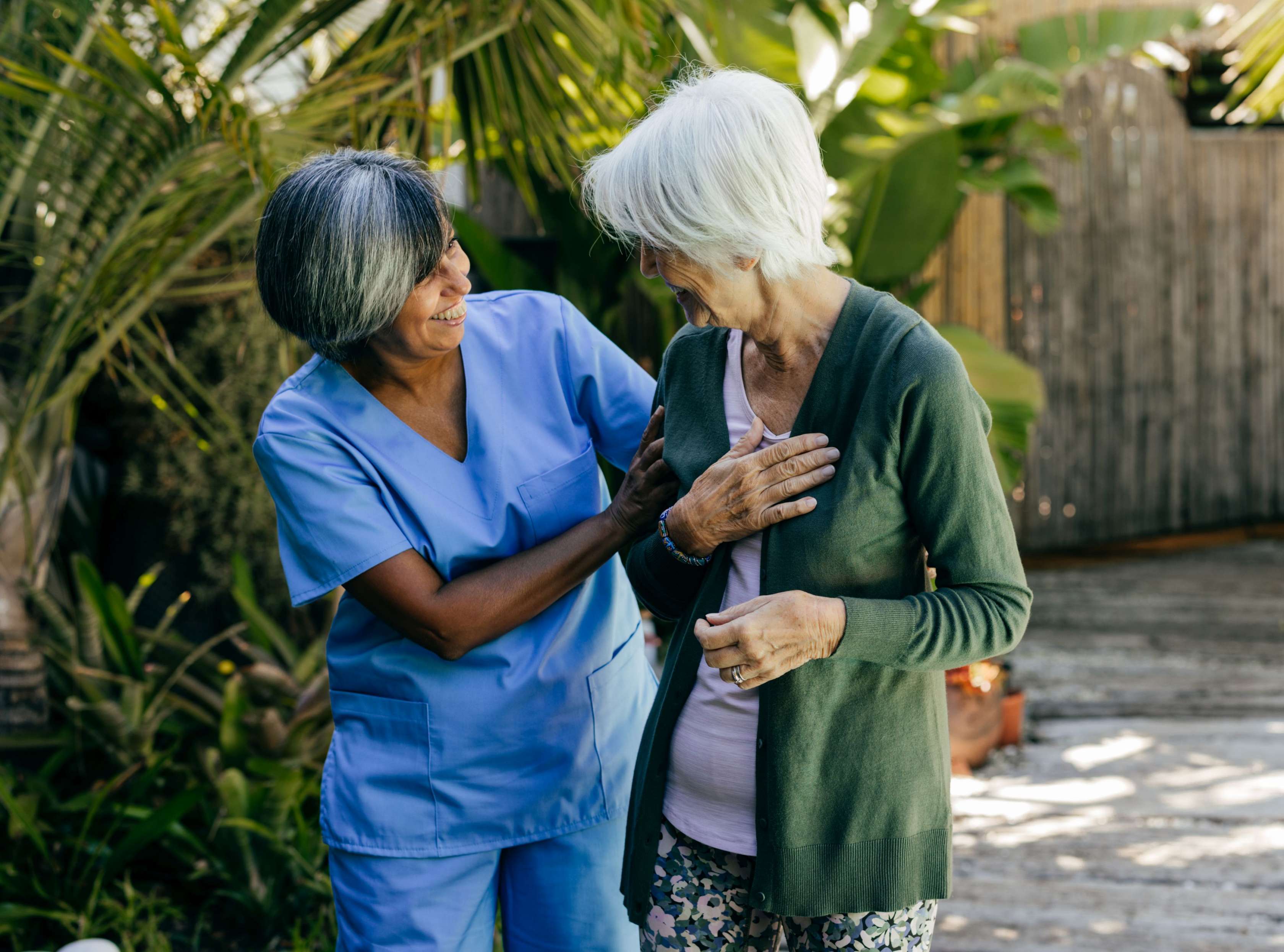 Pflege im Ausland: Eine weißhaarige Seniorin wird von einer Pflegerin in blauem Kasack beim Spazieren durch einen Palmengarten begleitet.