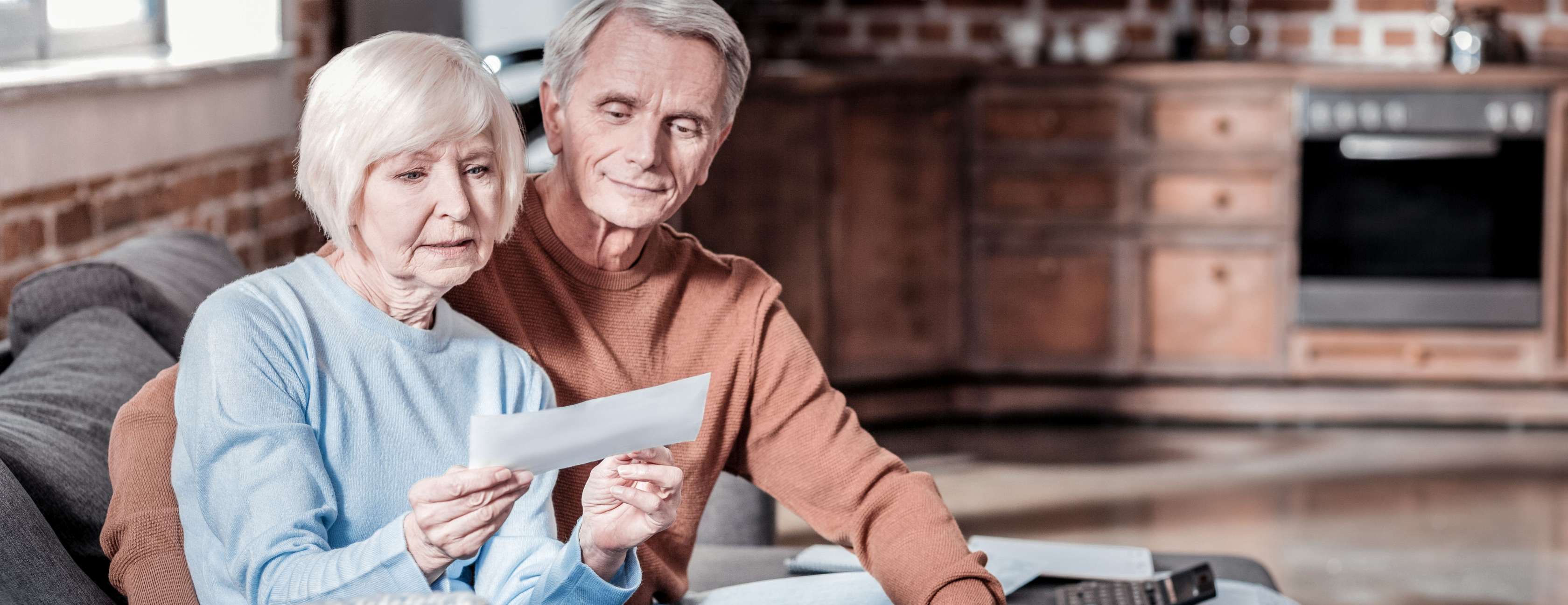 Älteres Paar auf einem Sofa schaut gemeinsam ein Dokument zu Pflegeheim-Kosten an.