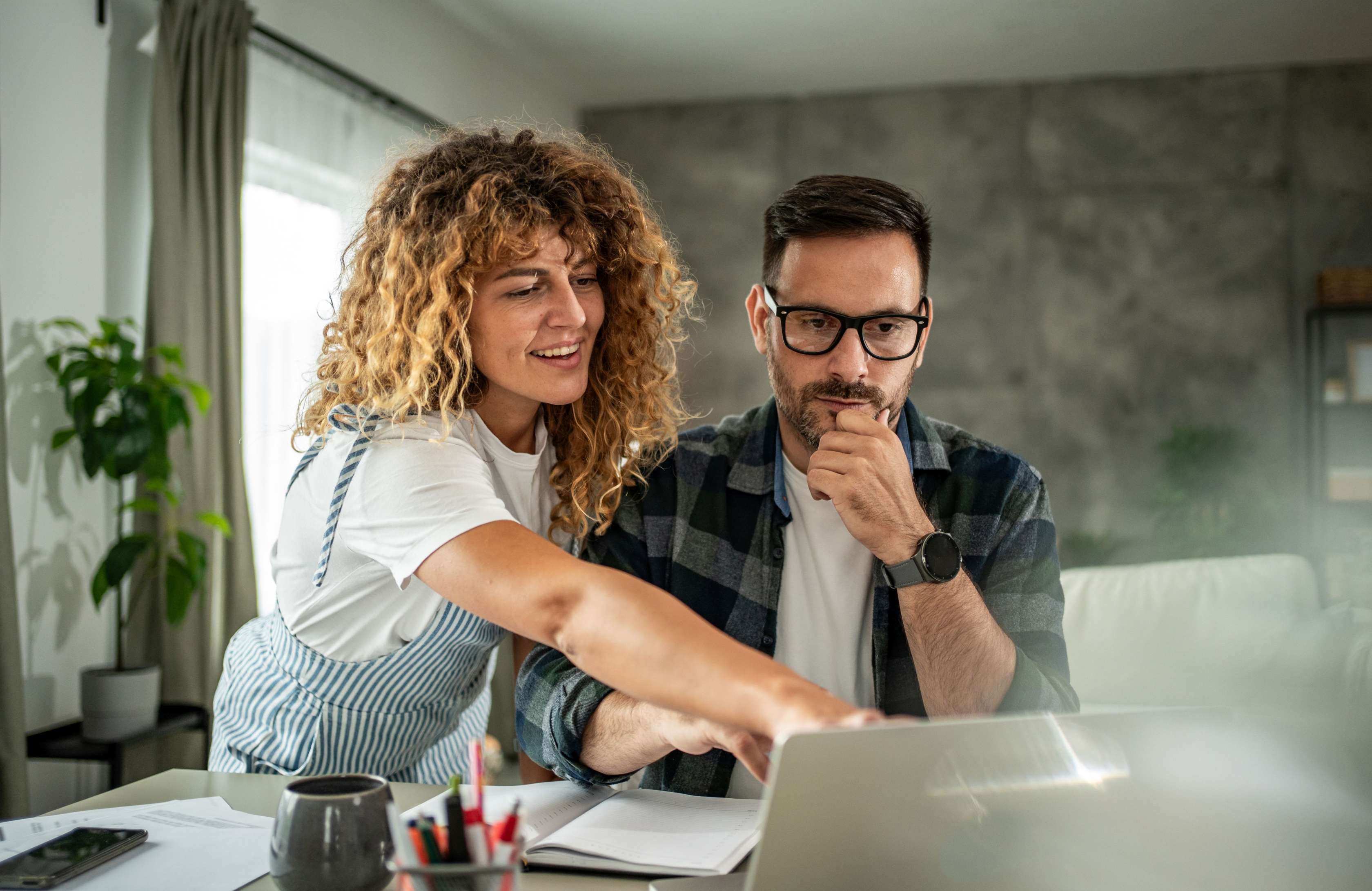 Eine Frau und ein Mann schauen am Schreibtisch gemeinsam auf einen Tablet. Die Frau zeigt stehend auf den Bildschirm. Vor dem sitzenden Mann liegt auf dem Tisch liegt ein aufgeschlagenes Notizbuch..