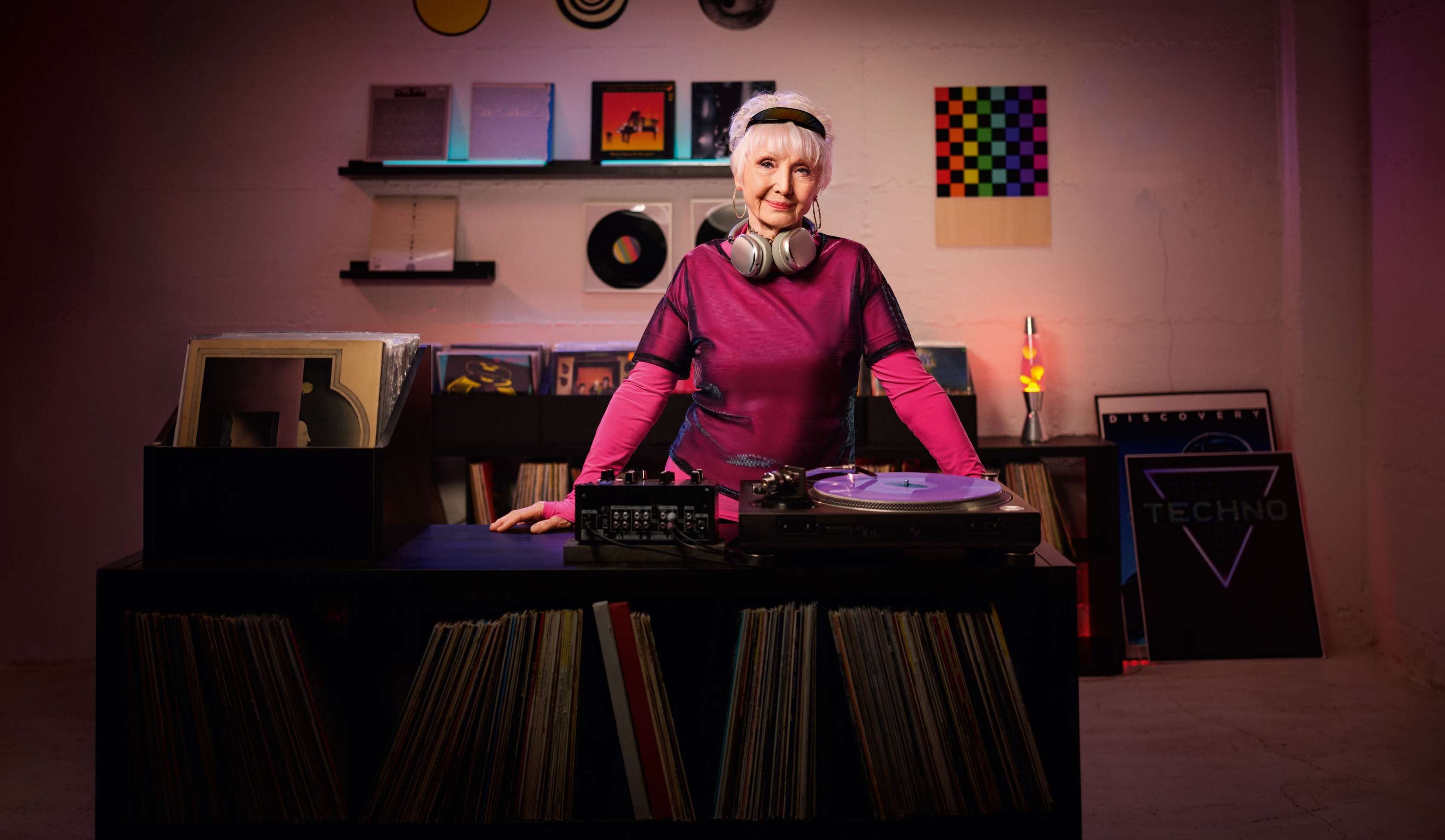 Allianz Pflegezusatzversicherung: Eine ältere Dame in pinker Kleidung steht hinter einem DJ-Pult