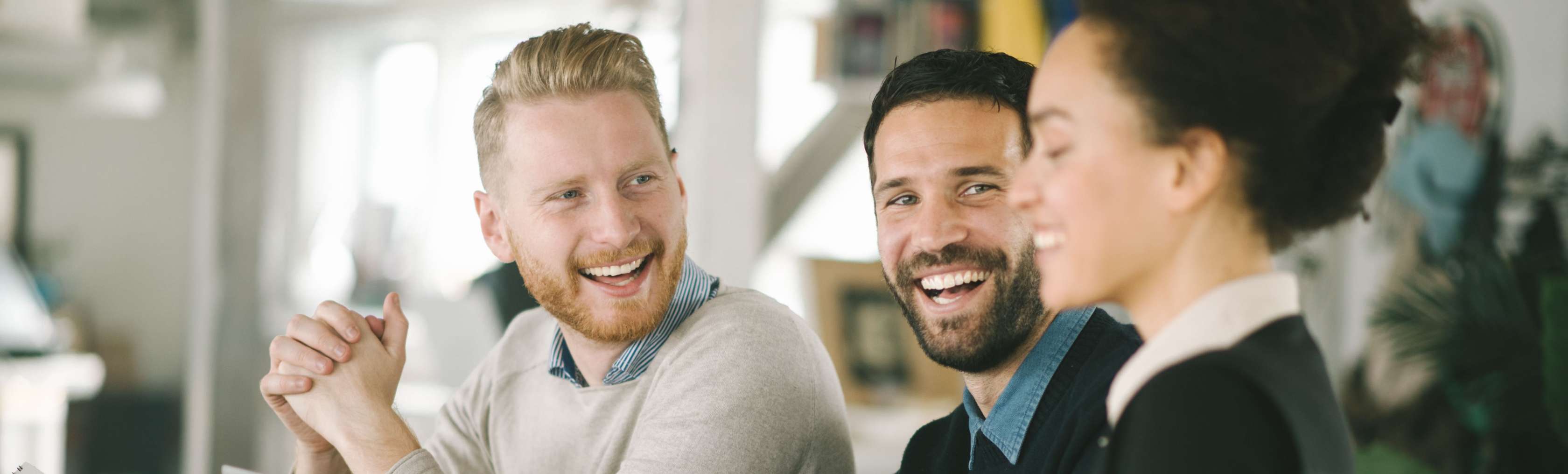 Allianz Private Kranken­versicherung für Angestellte: Kolleg:innen sitzen lachend mit Laptop und Kaffee an einem Tisch.