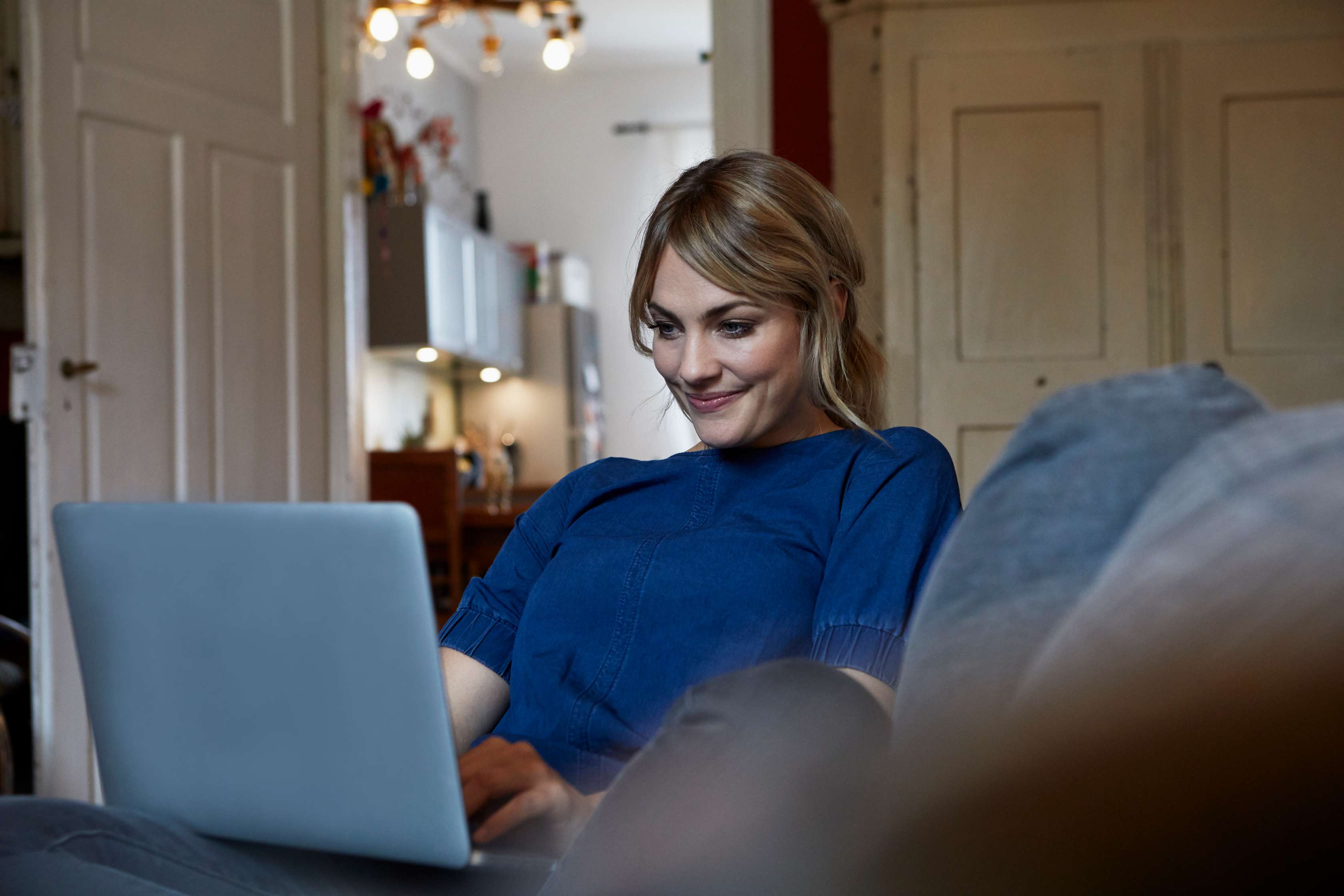 Junge Frau sitzt lächelnd mit Laptop auf dem Schoß auf dem Sofa