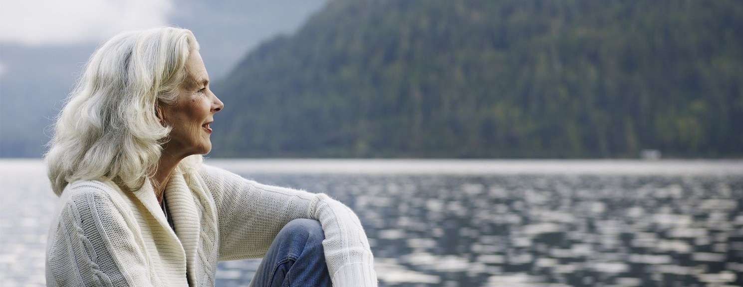 Ältere Frau sitzt auf Steg am See und blickt zuversichtlich und glücklich