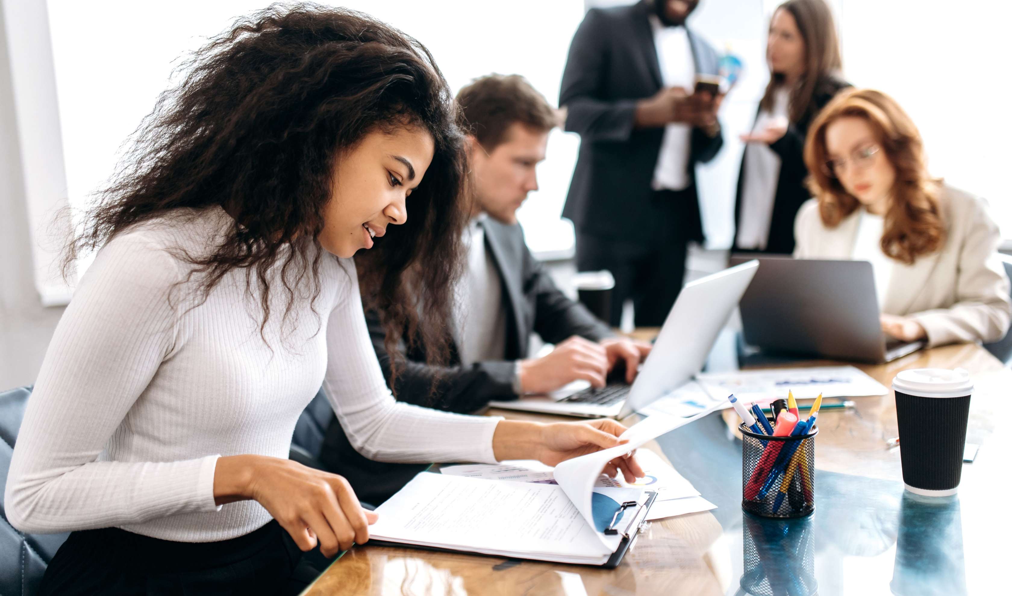 Private Krankenversicherung: Eine junge Frau blickt in einen aufgeschlagenen Block an einem Konferenztisch. Im Hintergrund sieht man weitere Personen vor Laptos sitzen oder im Gespräch.