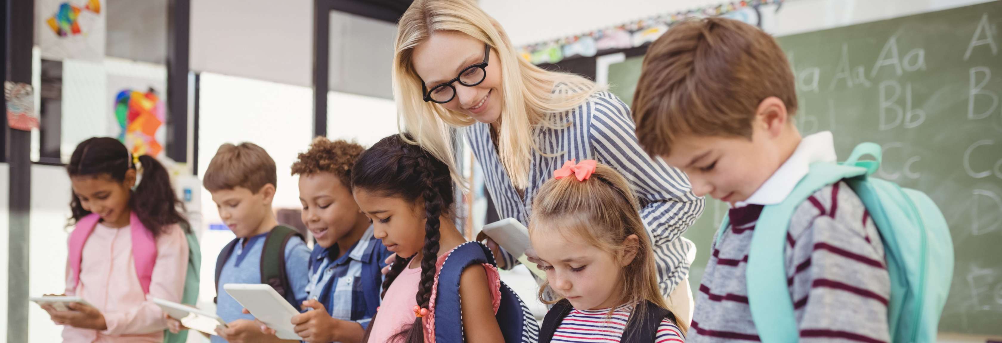 Junge Lehrerin mit blonden Haaren und Brille beugt sich lächelnd über eine Reihe von Schülern mit Notepads