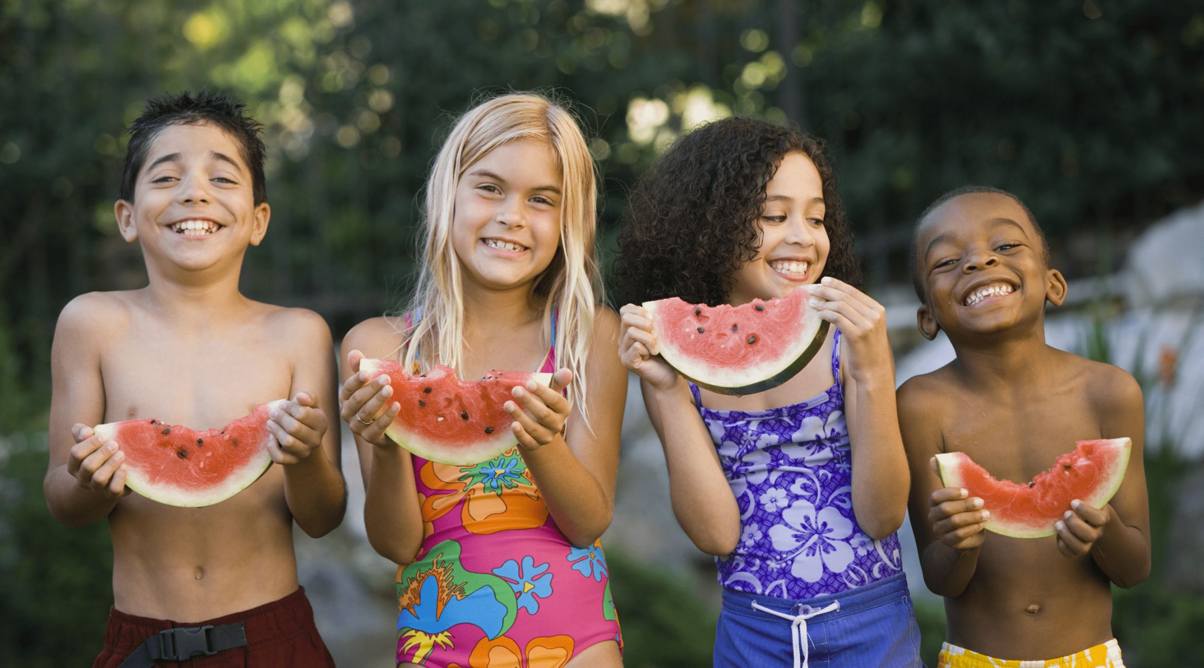 Vier Kinder stehen nebeneinander, freuen sich und essen jeder ein Stück Wassermelone